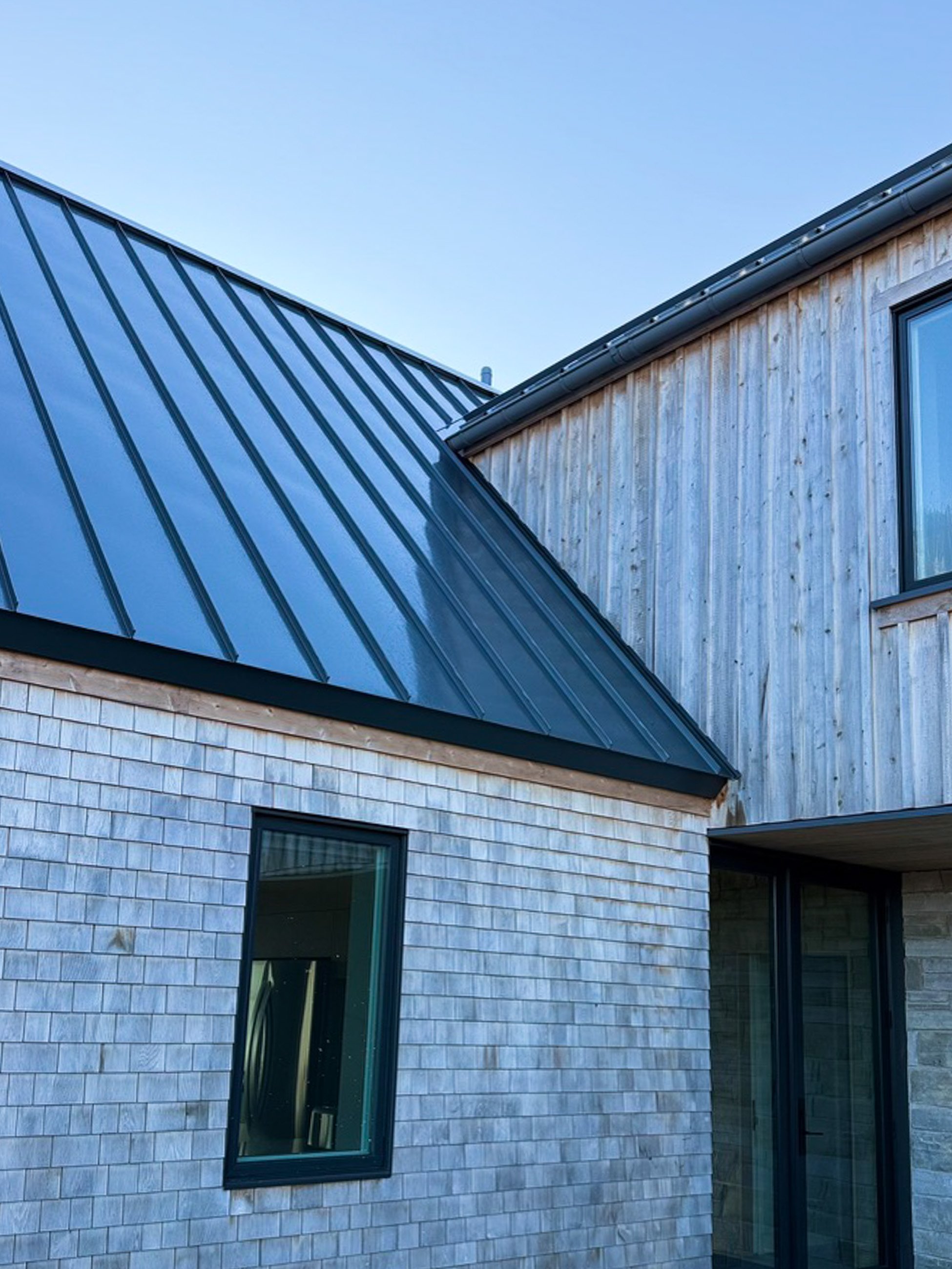Close-up of a modern house with a pitched metal roof, wooden siding, and a window, under a clear blue sky.