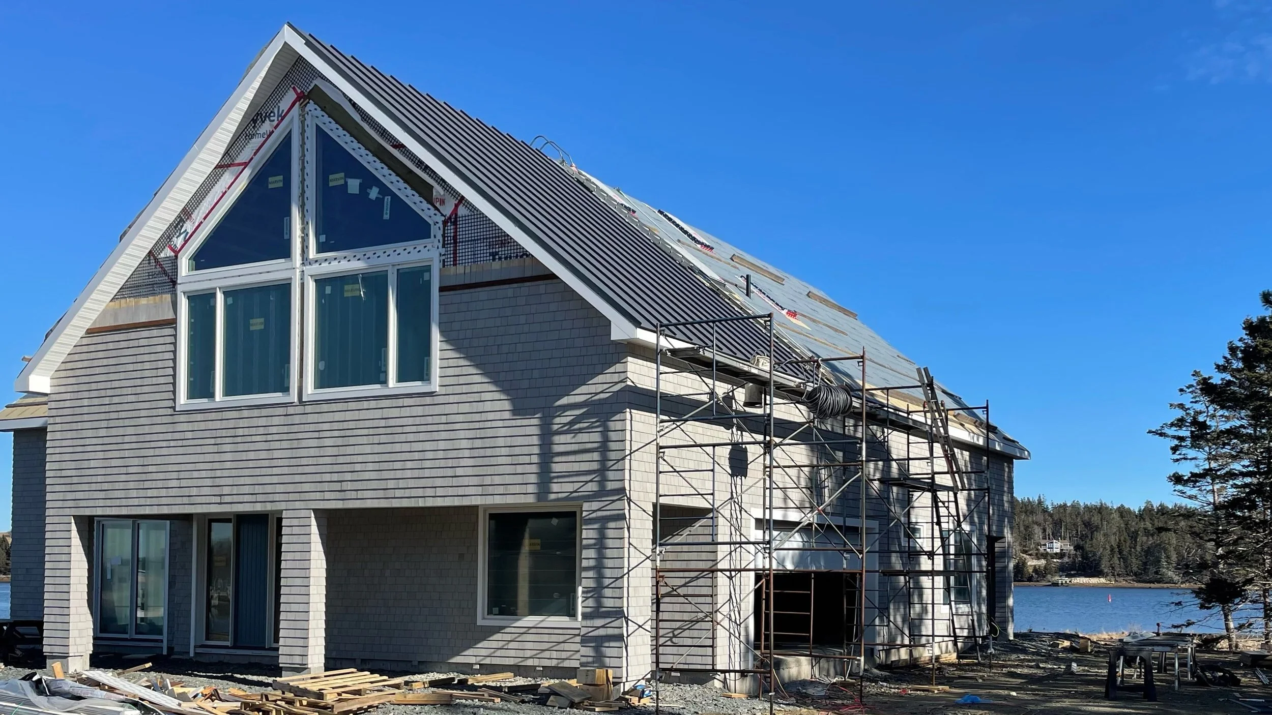 A house under construction by a body of water with scaffolding around it and a steep roof with roofing materials being installed.