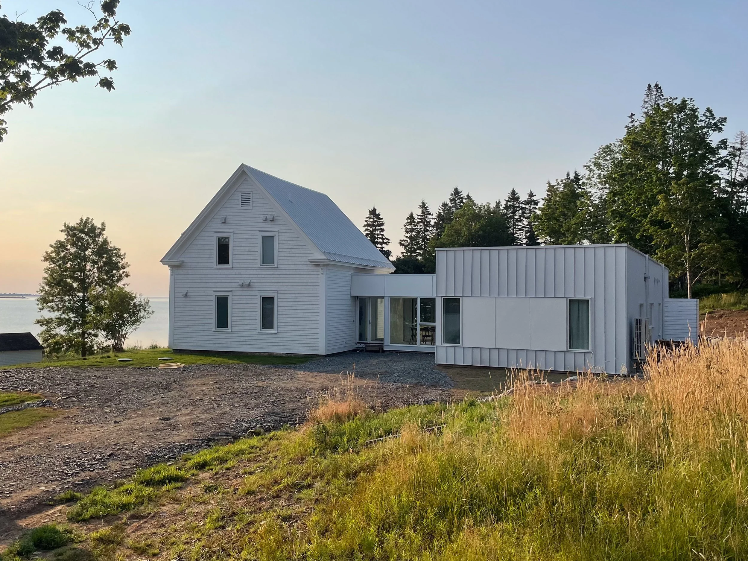 Modern white house with two sections, situated near water, surrounded by trees and tall grass, during sunset.