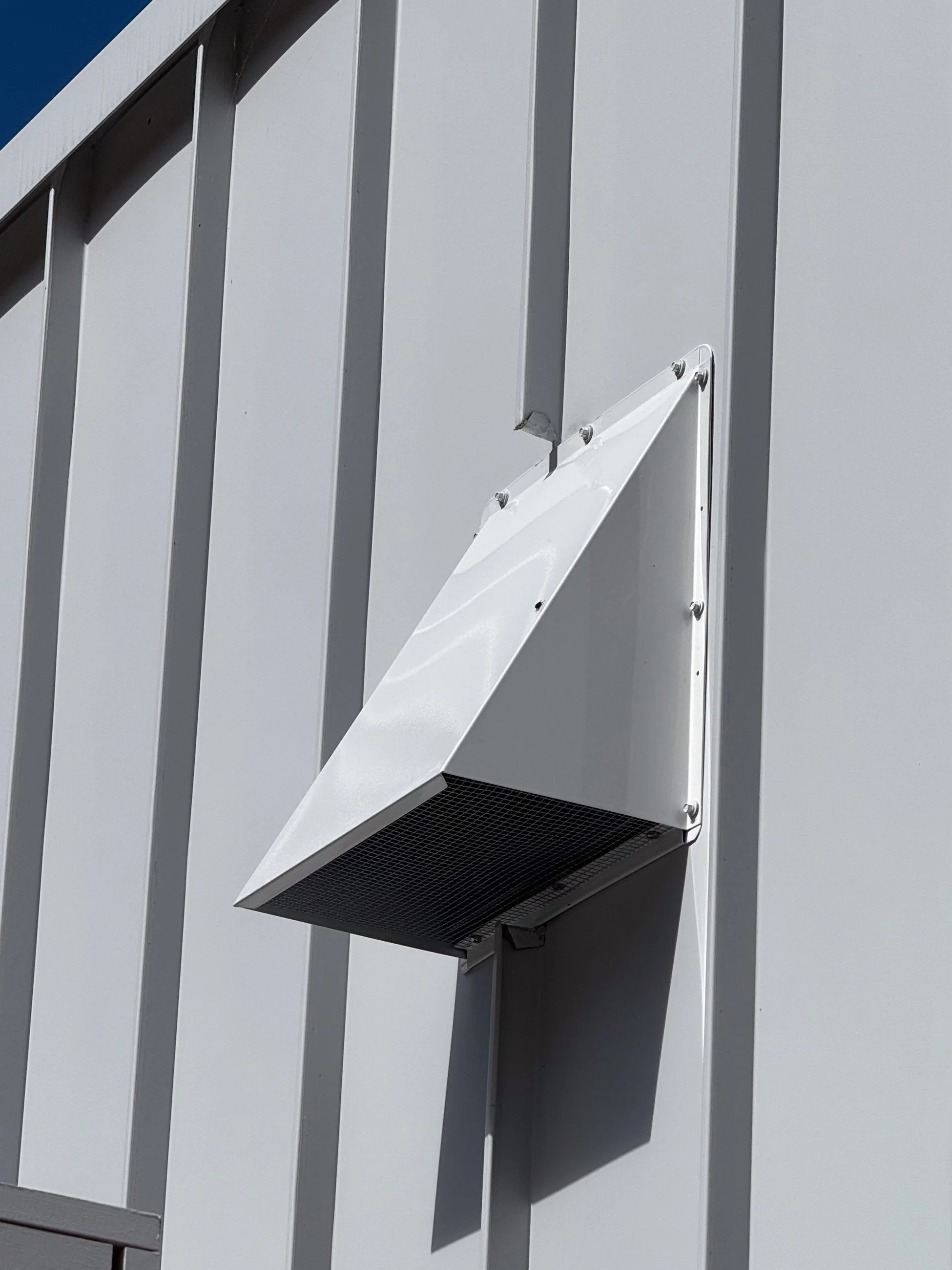 Exterior view of a metal building wall with a mounted white exhaust vent or air intake with a protective grille, cast in sunlight.