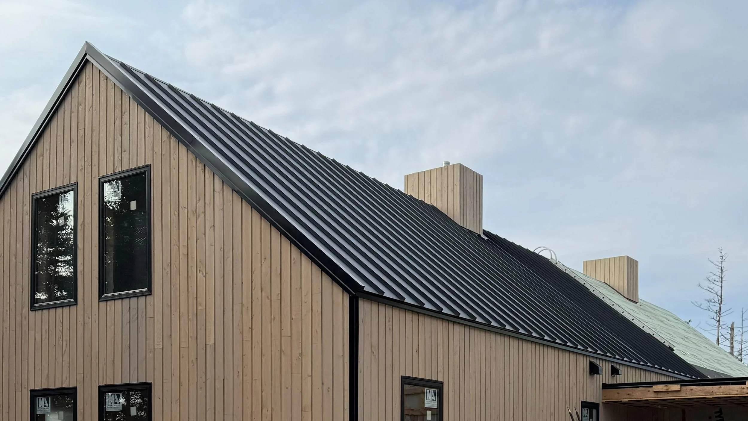 Close-up of a modern house with a black metal roof and light wood siding, with multiple rectangular windows.