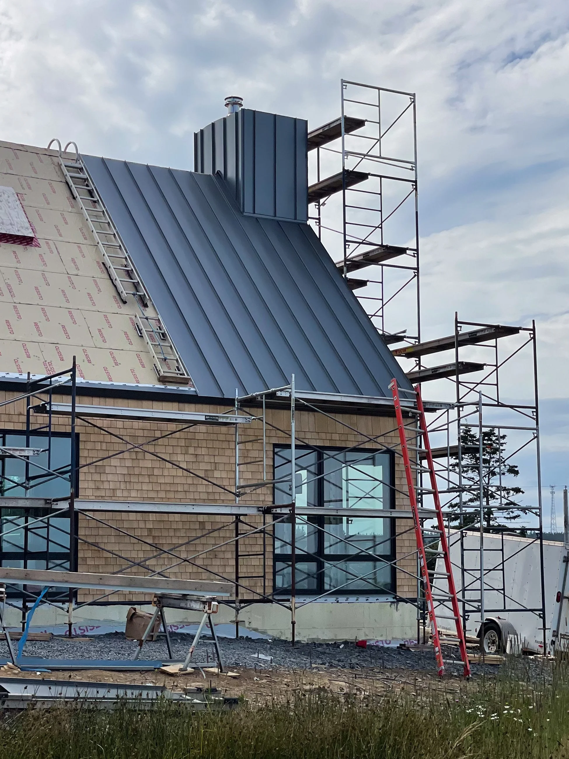 Construction site with scaffolding around a building that has a diagonal metal roof and large windows, with part of the roof under construction, tools, and equipment visible.