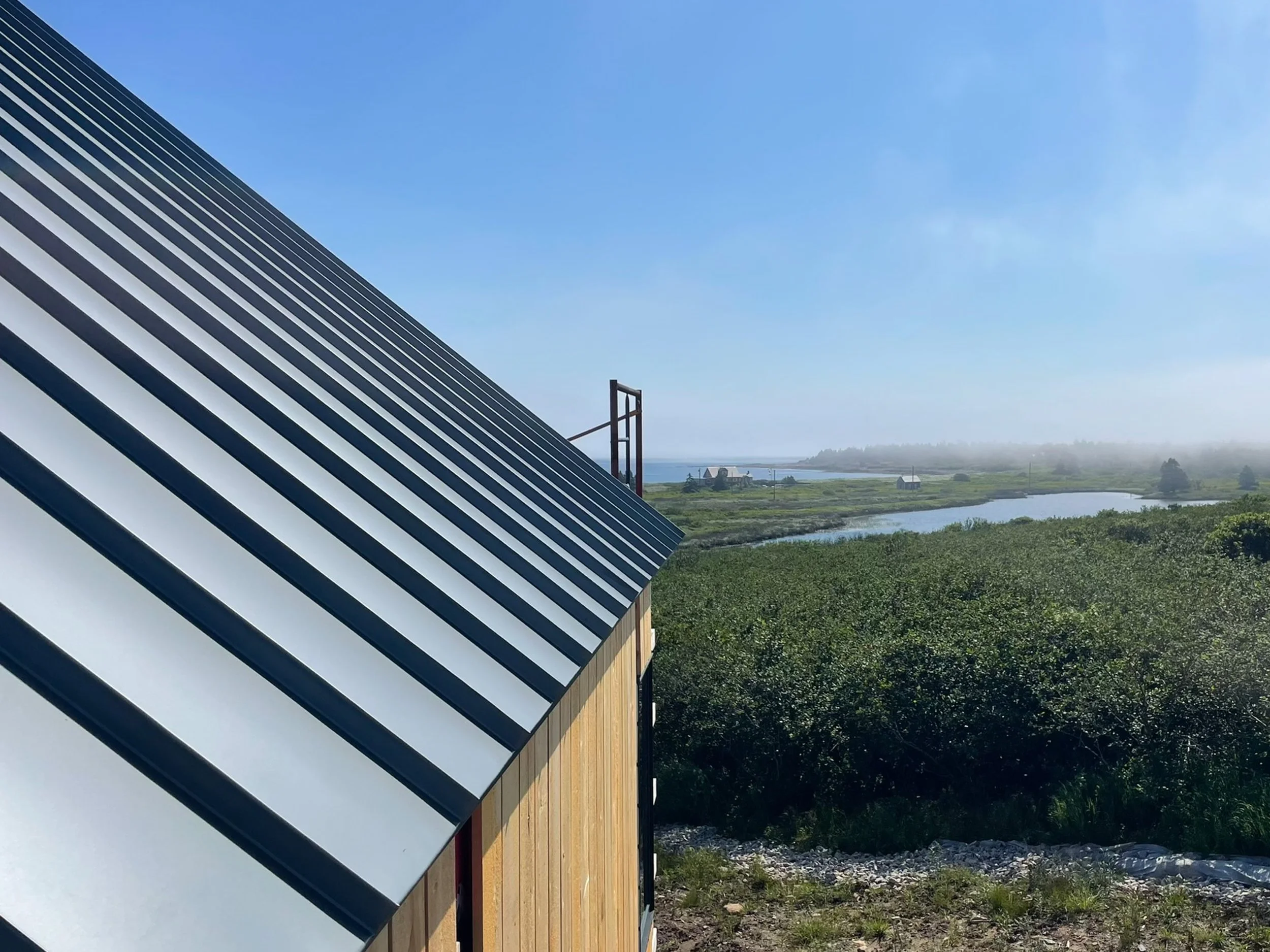 A house with a metal roof on a green landscape near water, under a clear blue sky with some fog in the distance.