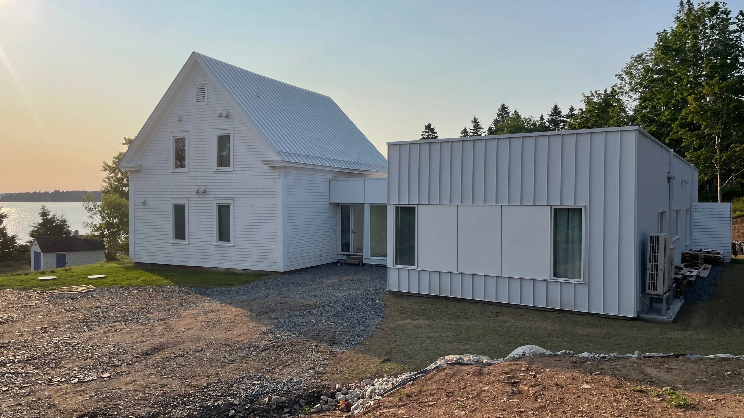 Two modern houses are located near a body of water with trees in the background. One house is a traditional white wooden structure, and the other is a contemporary white metal house.