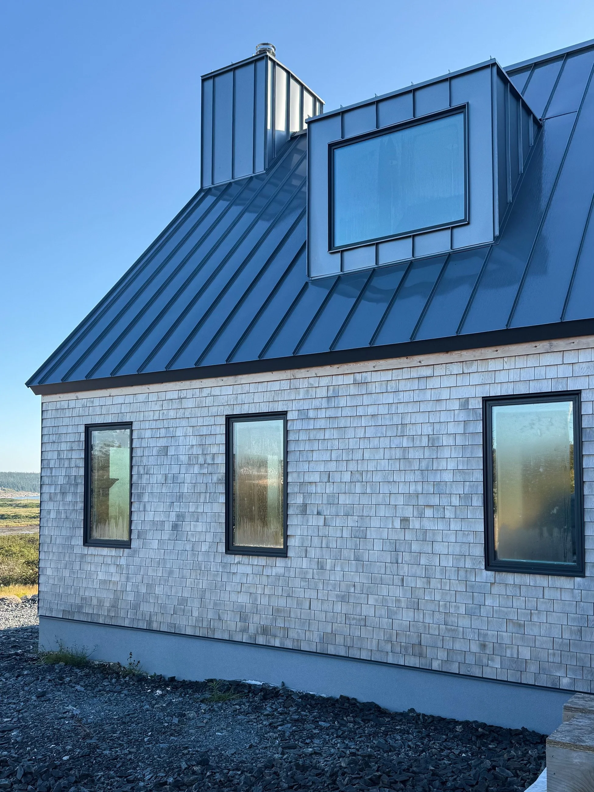 A building with a dark metal roof, light-colored shingle siding, and three rectangular windows. There are two roof access structures, one is a dormer and the other appears to be an attic ventilation. The scene is outdoors with a clear blue sky.