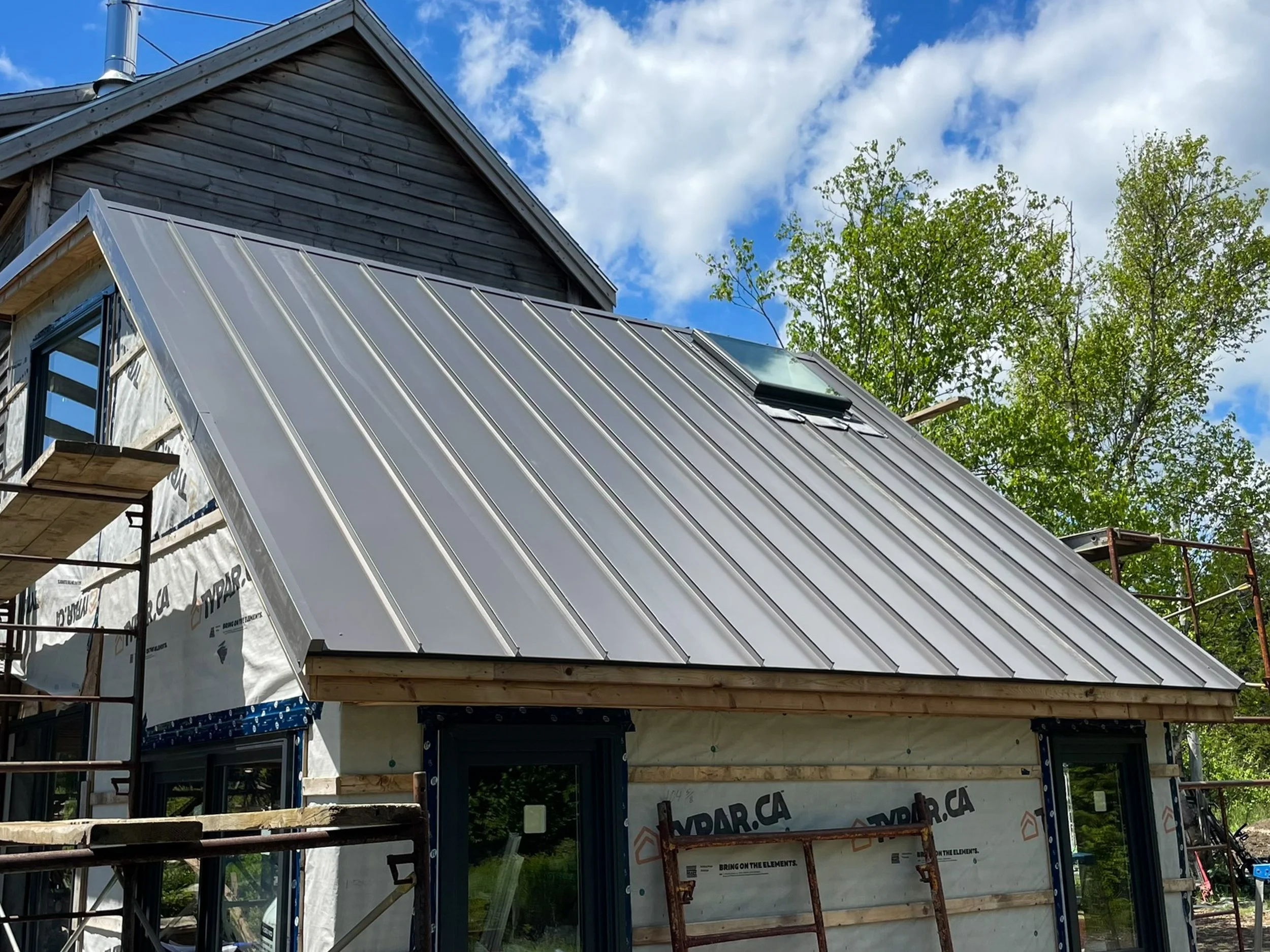 Under construction house with a metal roof, window, and surrounding scaffolding, under a partly cloudy sky with green trees in the background.