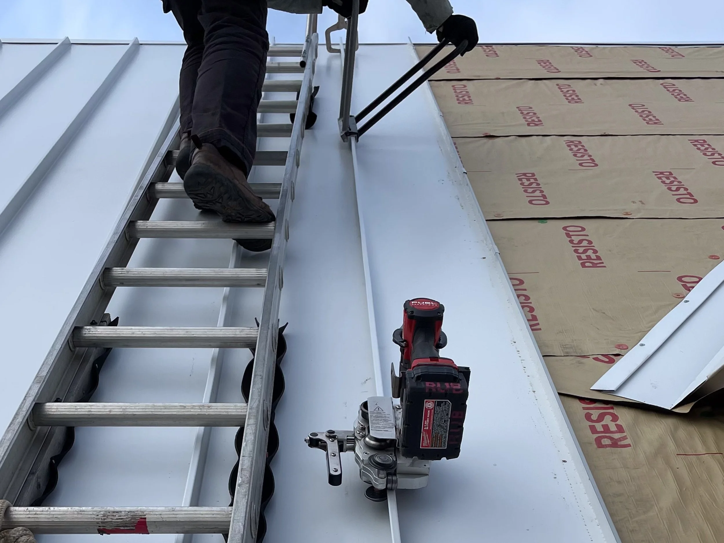 Person installing or repairing siding on a building, using a ladder, a cordless drill, and safety brackets on the siding.