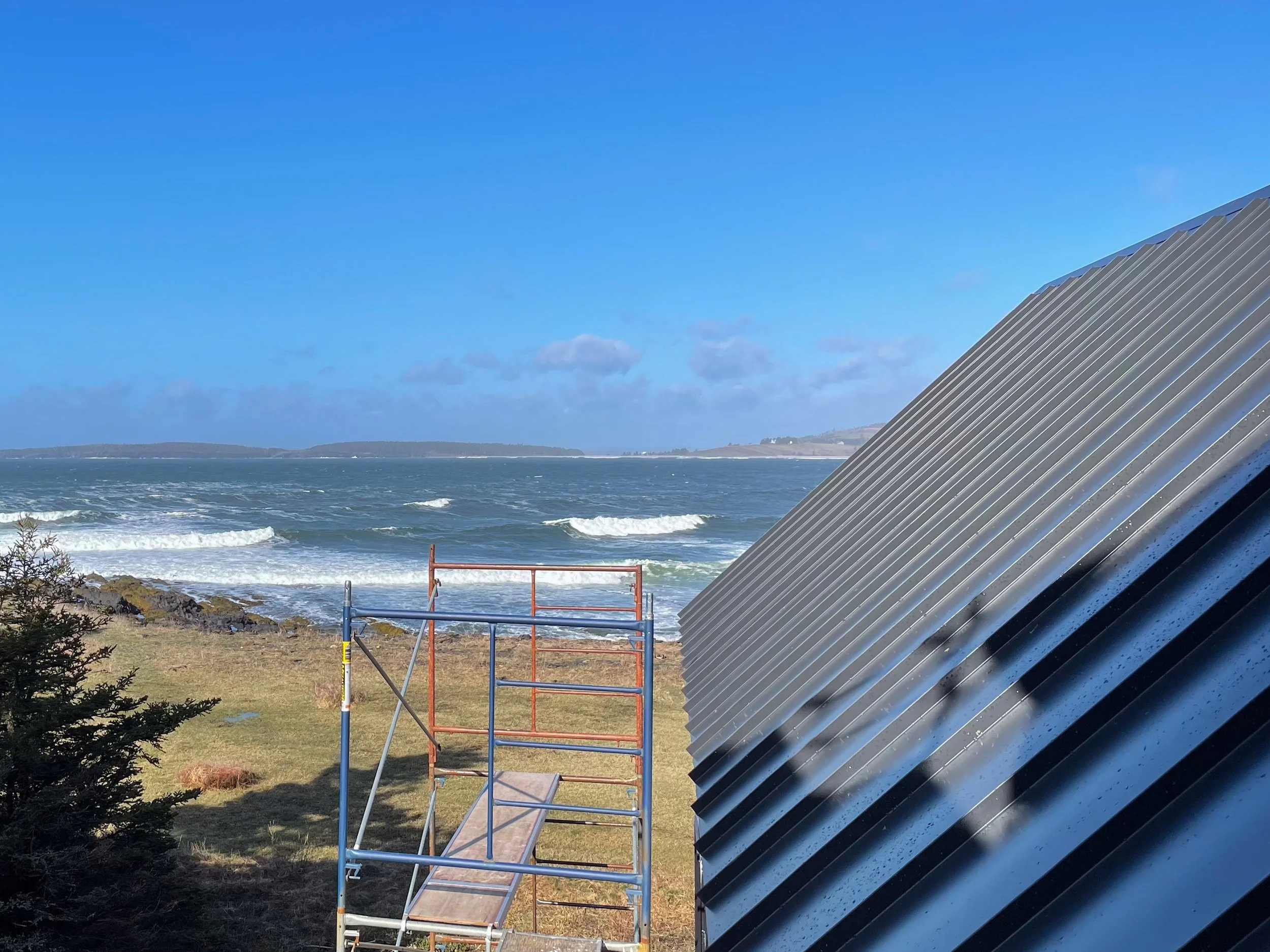 View of the ocean with waves and a distant horizon, a blue sky with some clouds, a grassy area with a small bush, a metal scaffold, and part of a large metal roof in the foreground.