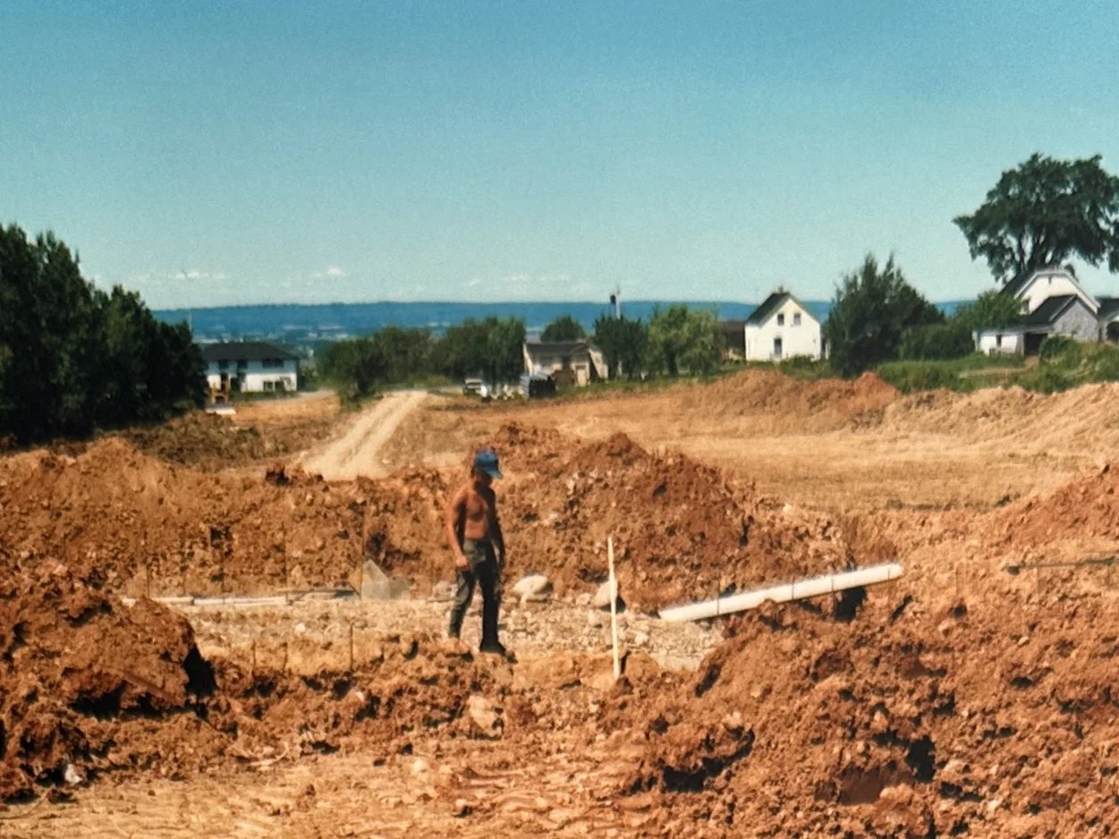 A man without a shirt, wearing a blue hat, standing on a construction site with dirt piles and a pipe, in a rural area with houses and trees in the background.