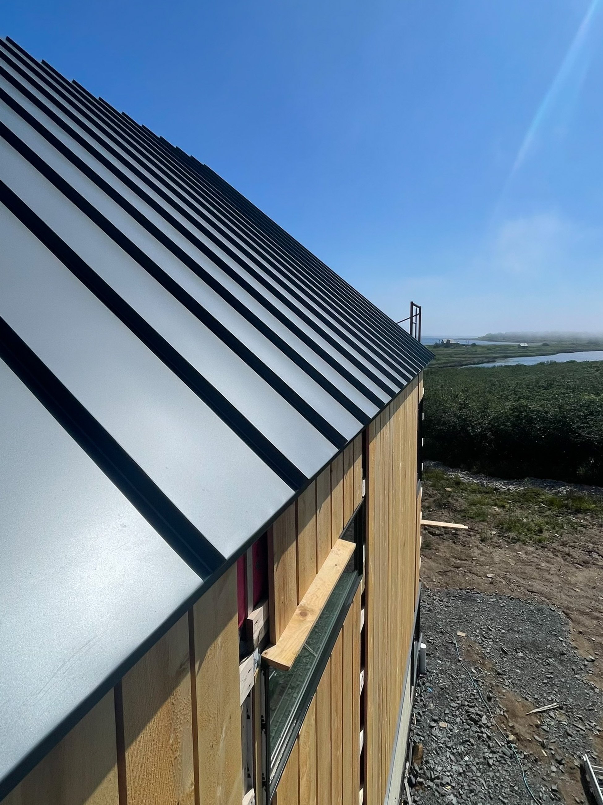 Side of a building with a metal roof, partial window, and wooden siding, set against a clear blue sky with some landscape in the distance.