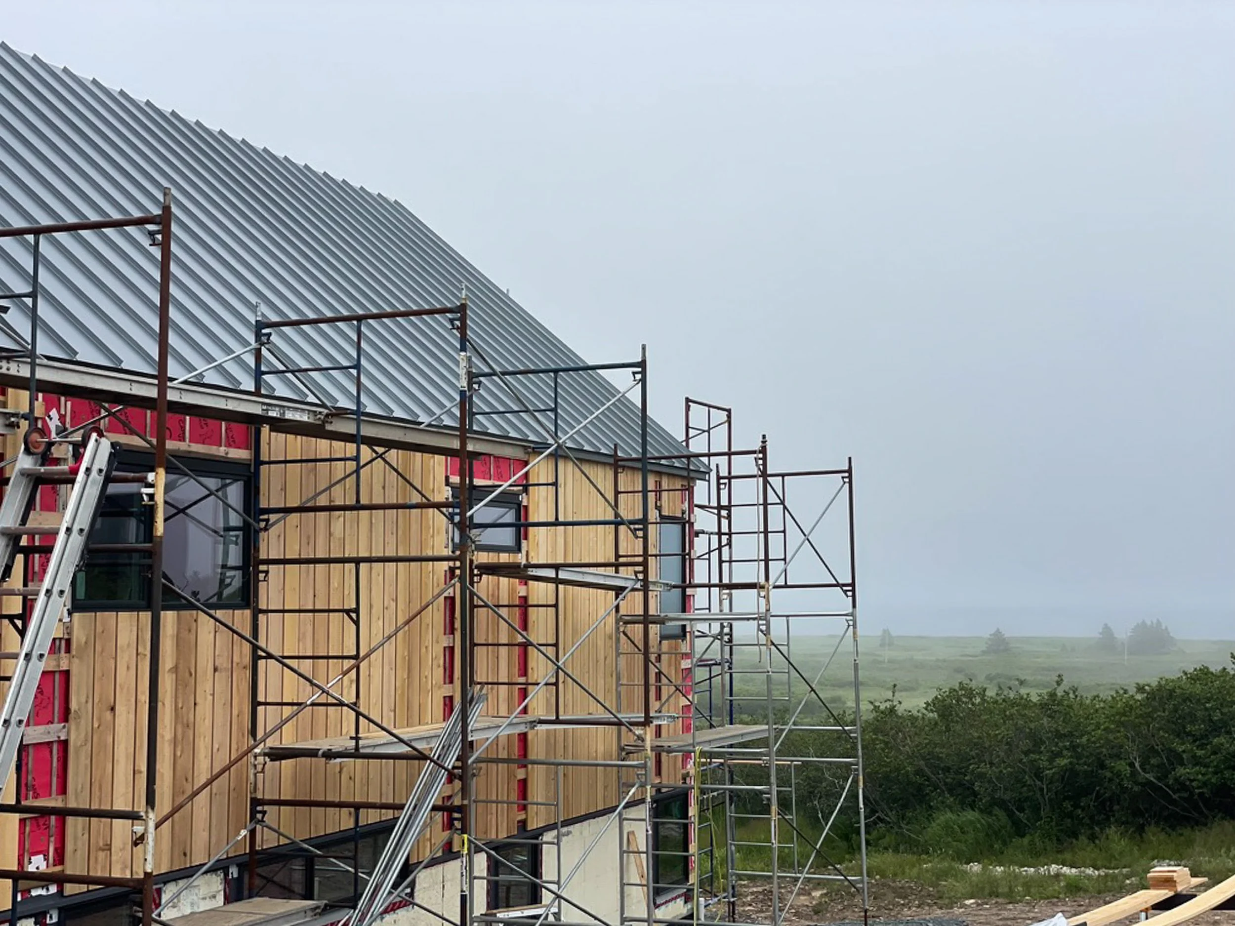 Construction of a new house with scaffolding around it, wooden exterior panels being installed, and a metal roof partially completed on a cloudy day with greenery in the background.