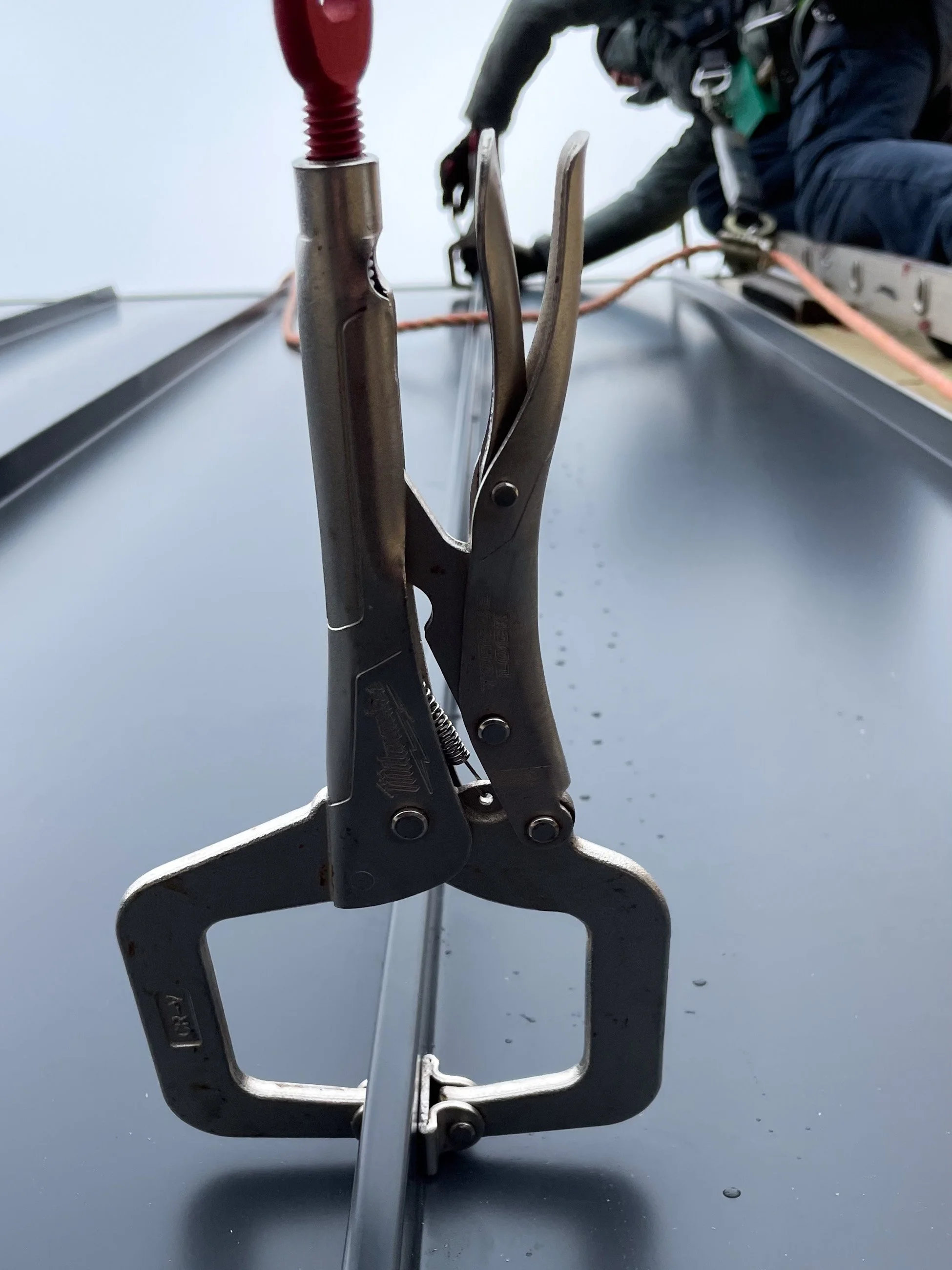 Close-up of a metal clamp device attached to a smooth gray surface, with construction workers in safety gear working in the background.