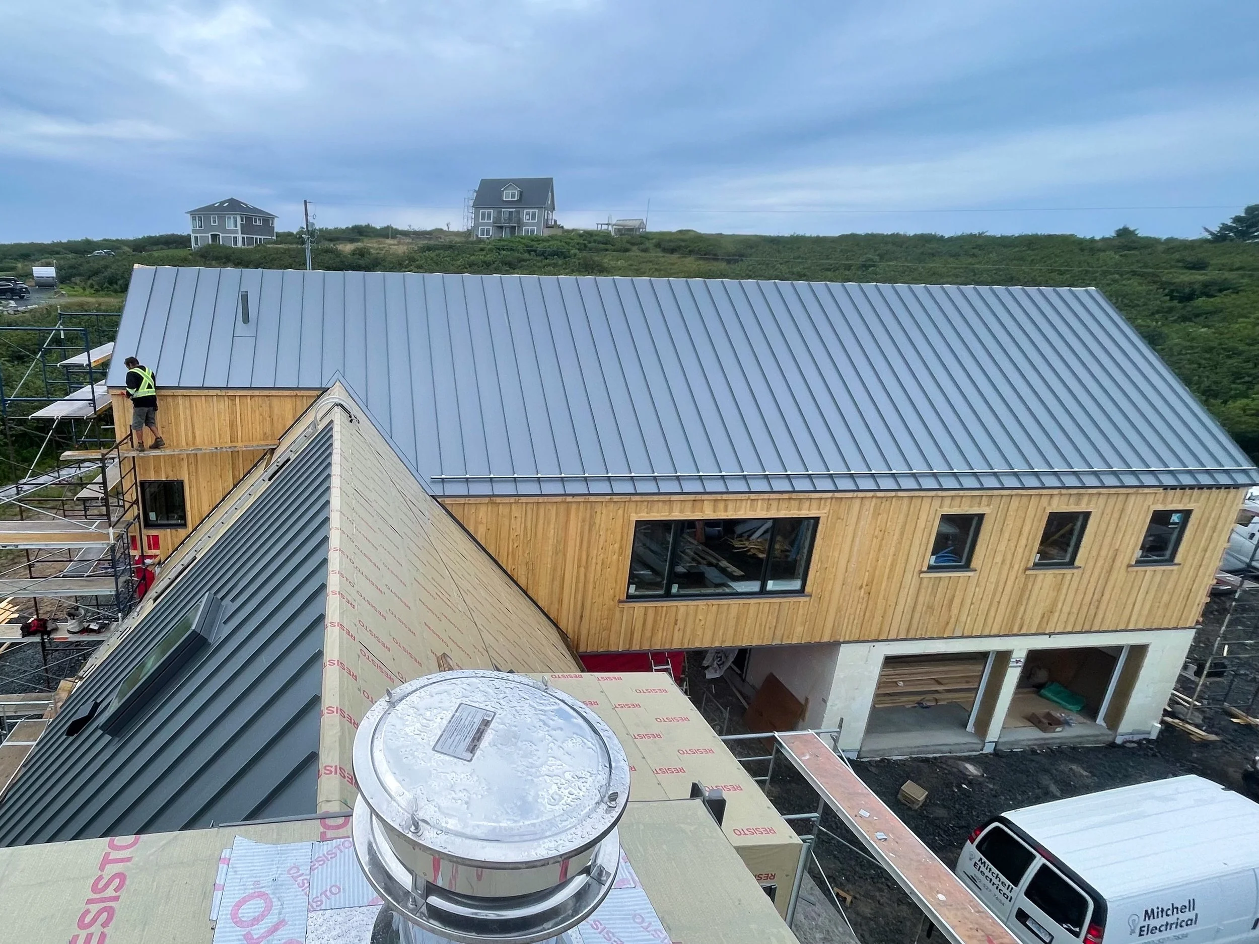 Under construction house with wooden siding and a blue metal roof, scaffolding on side, construction worker working, parked van outside, two houses in distance on hill.