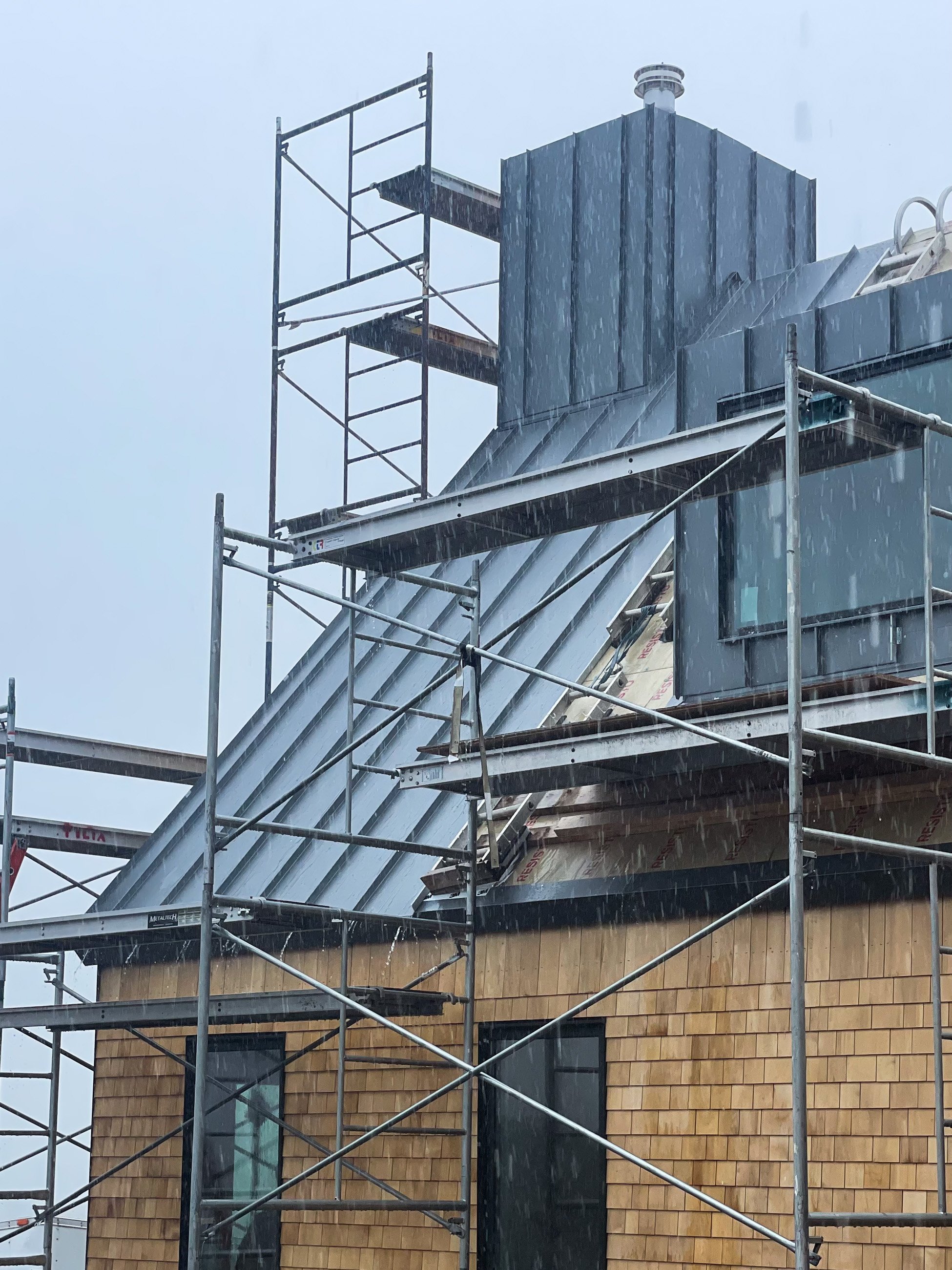 Construction scaffolding around a building with wooden siding, metal roof, and a chimney during a rainstorm.