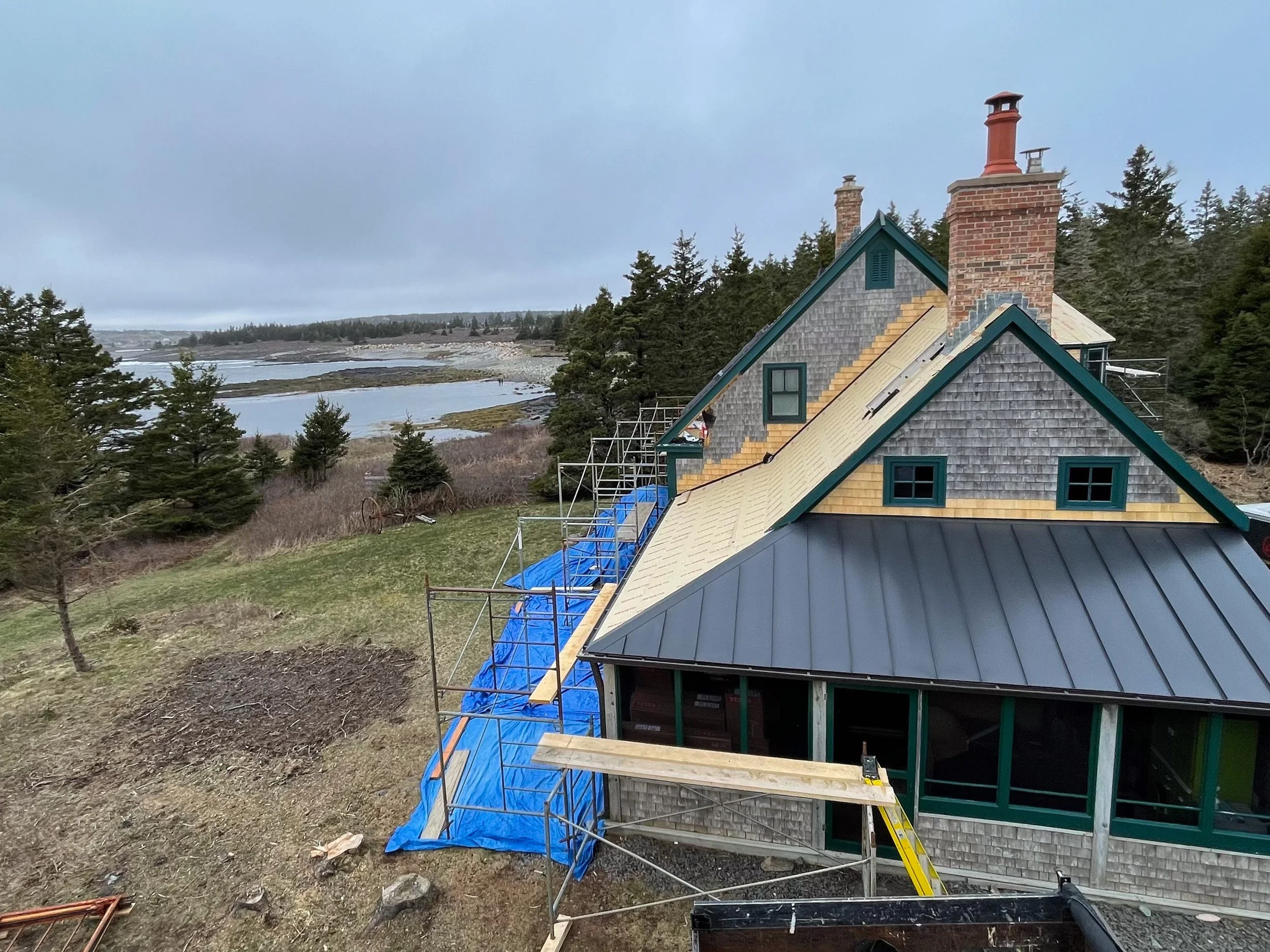 House under construction with a partially installed metal roof, scaffolding, and blue tarps, set in a landscape with trees and a body of water in the background.