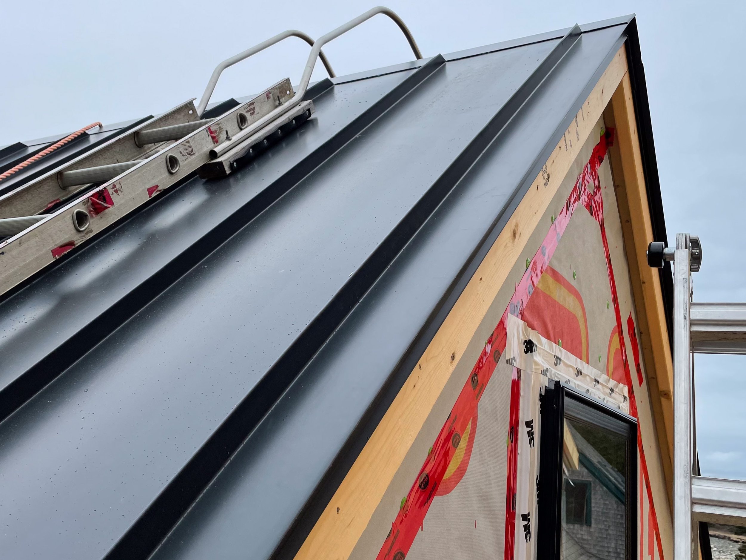 Close-up of a building under construction with a black metal roof, a ladder mounted on the roof, and window framing with red tape and weather-resistant material.