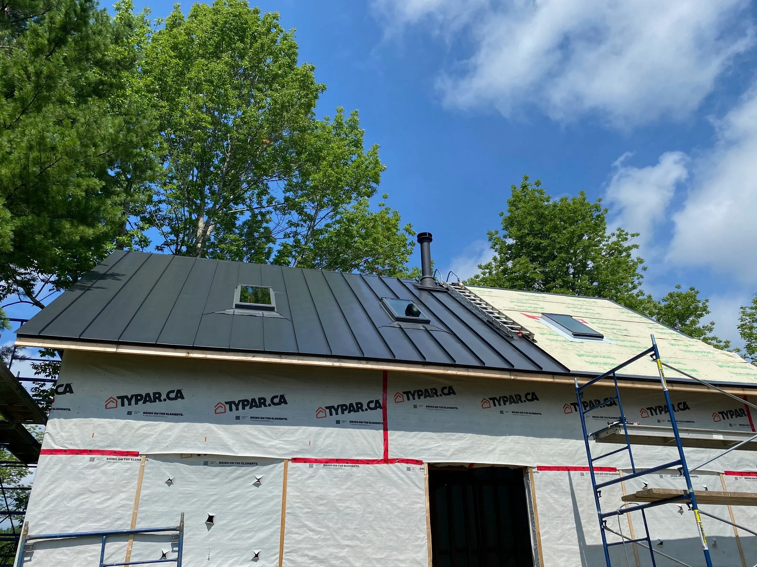 Under construction house with black metal roof, scaffolding, and green trees with blue sky and clouds in the background.