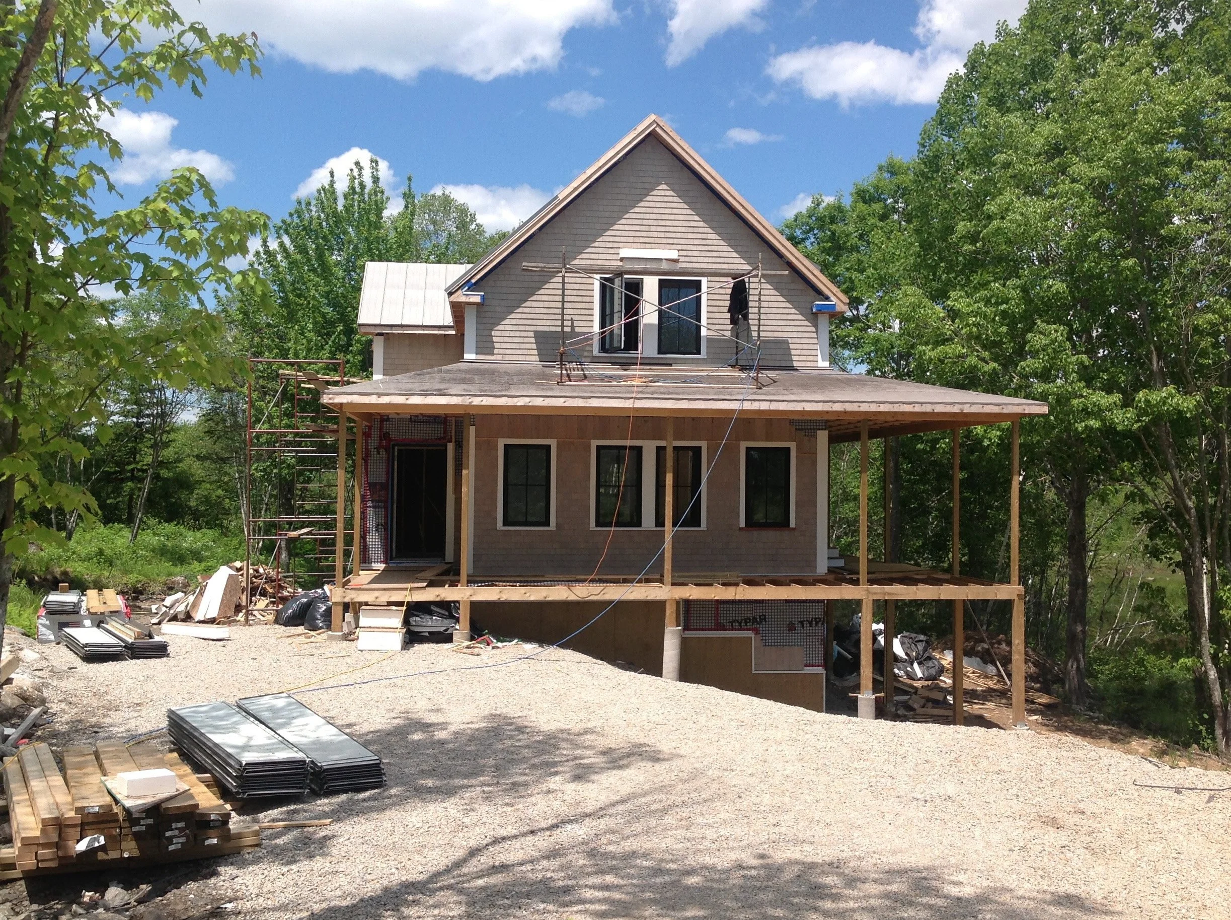 A two-story house under construction with scaffolding on the roof, surrounded by trees, and construction materials on the ground.