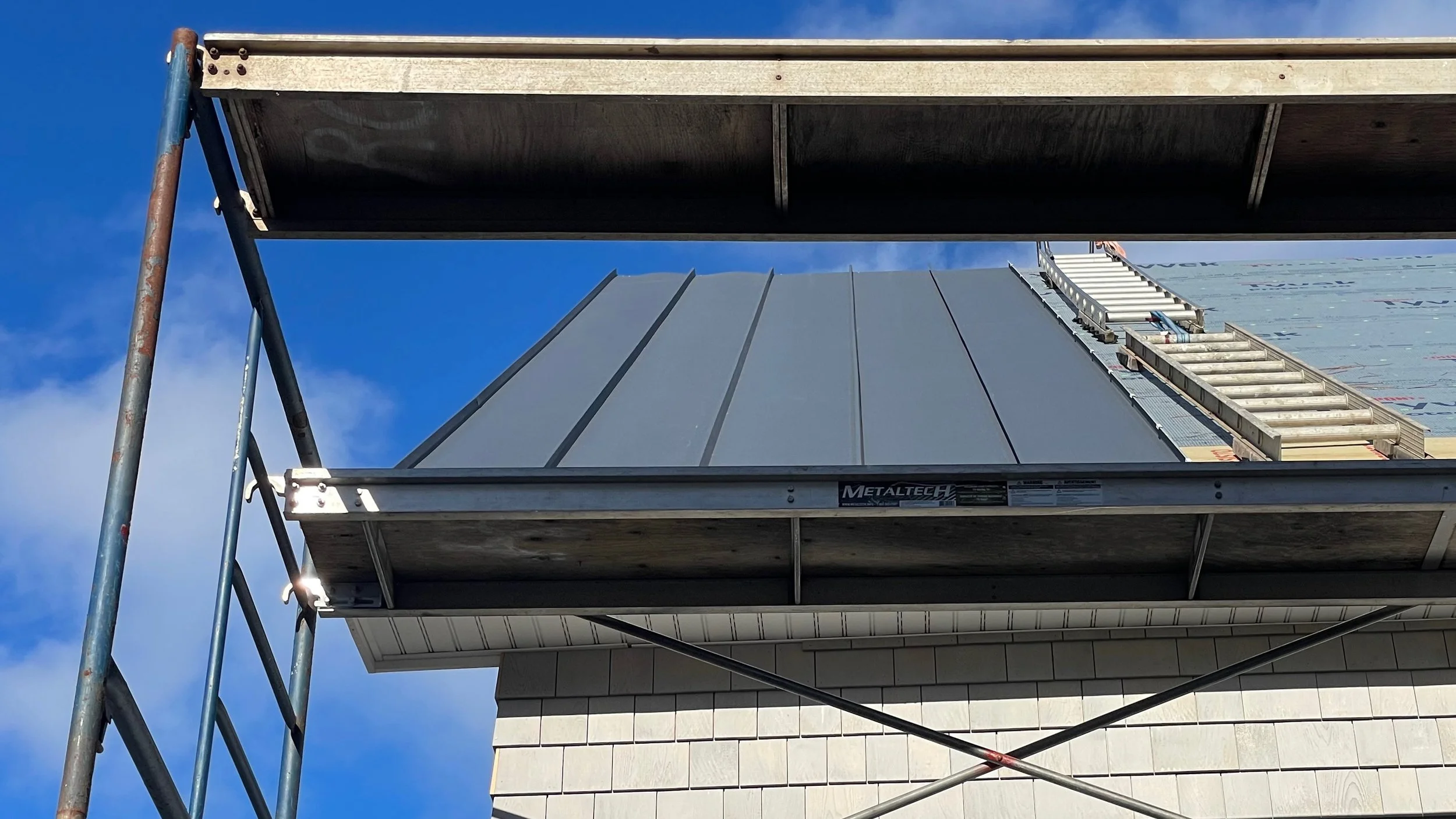 Construction scaffolding and roofing materials on a building under construction, with blue sky and some clouds in the background.