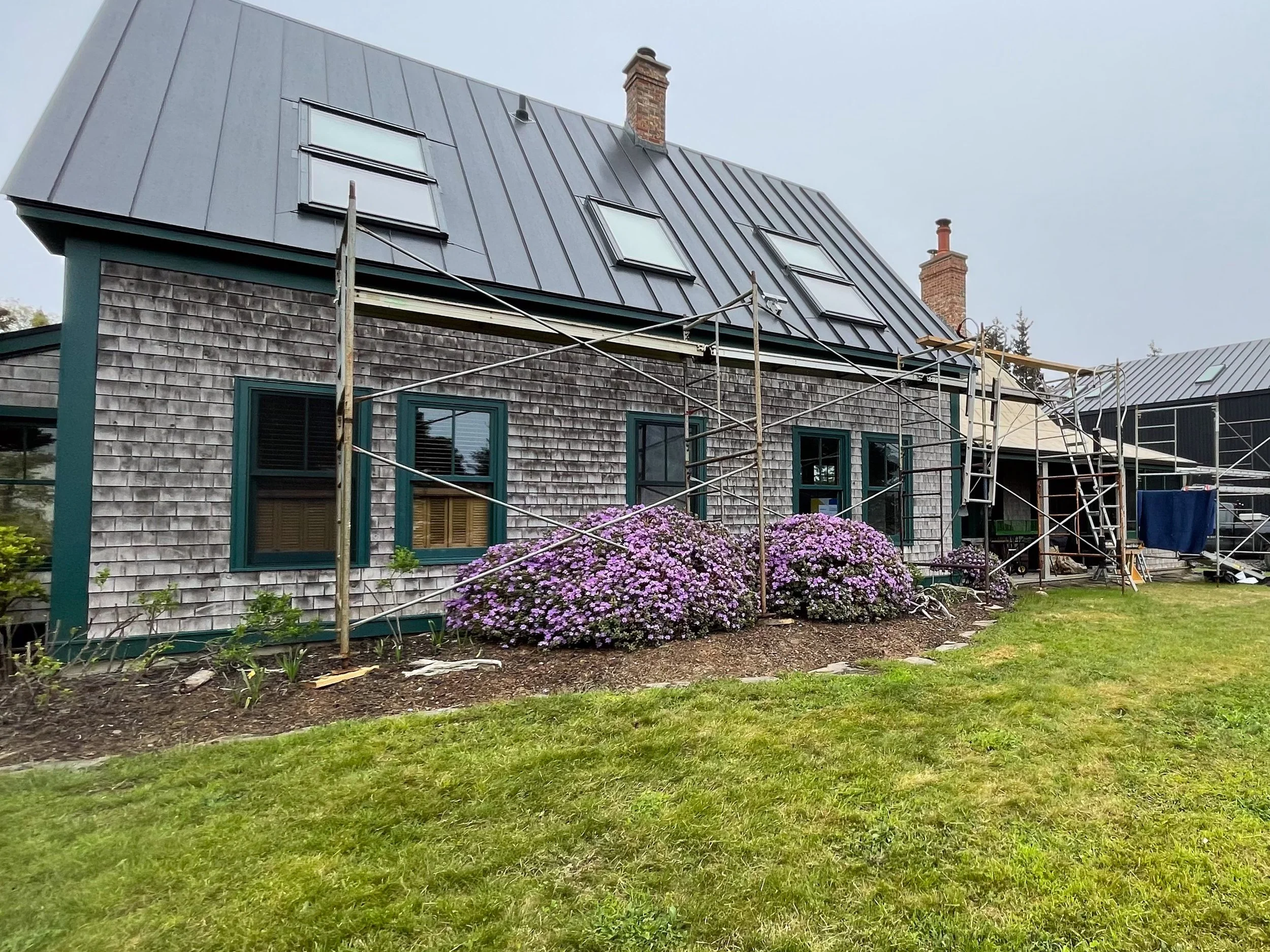 House under renovation with scaffolding, gray shingles, green window frames, purple flowers in front, and a metal roof.