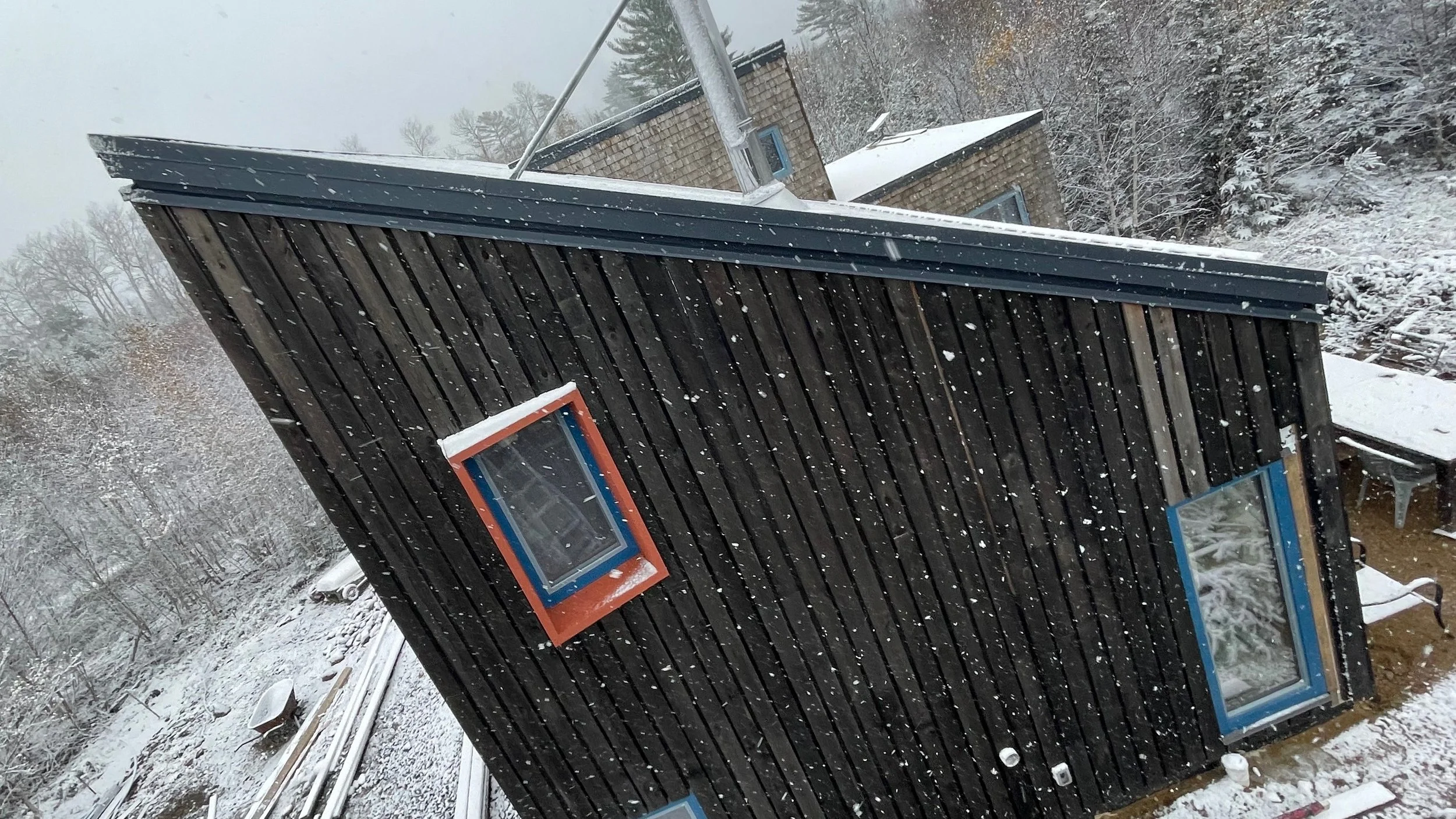A modern house with black wooden siding, red and blue-framed windows, surrounded by snow, with a snowy landscape and trees in the background.