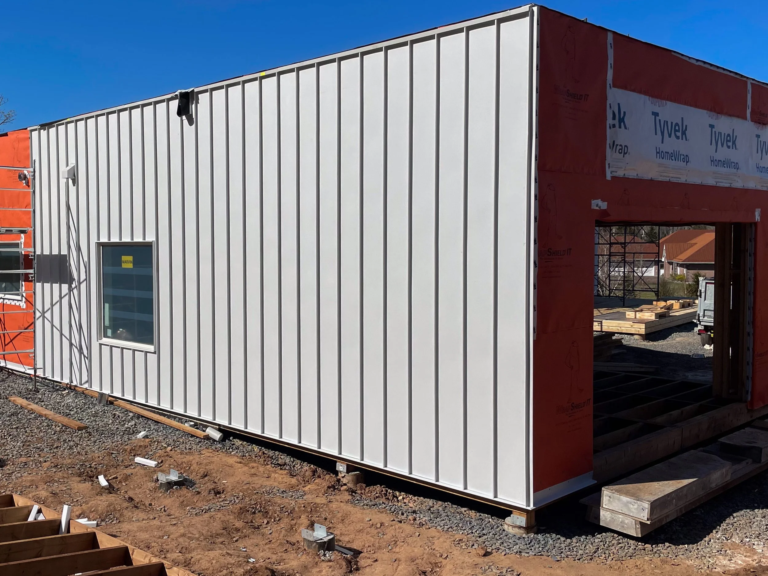 A building under construction with white metal siding on one side, a window, and an open section framed with red insulation material. Construction materials and scaffolding are visible around the site.