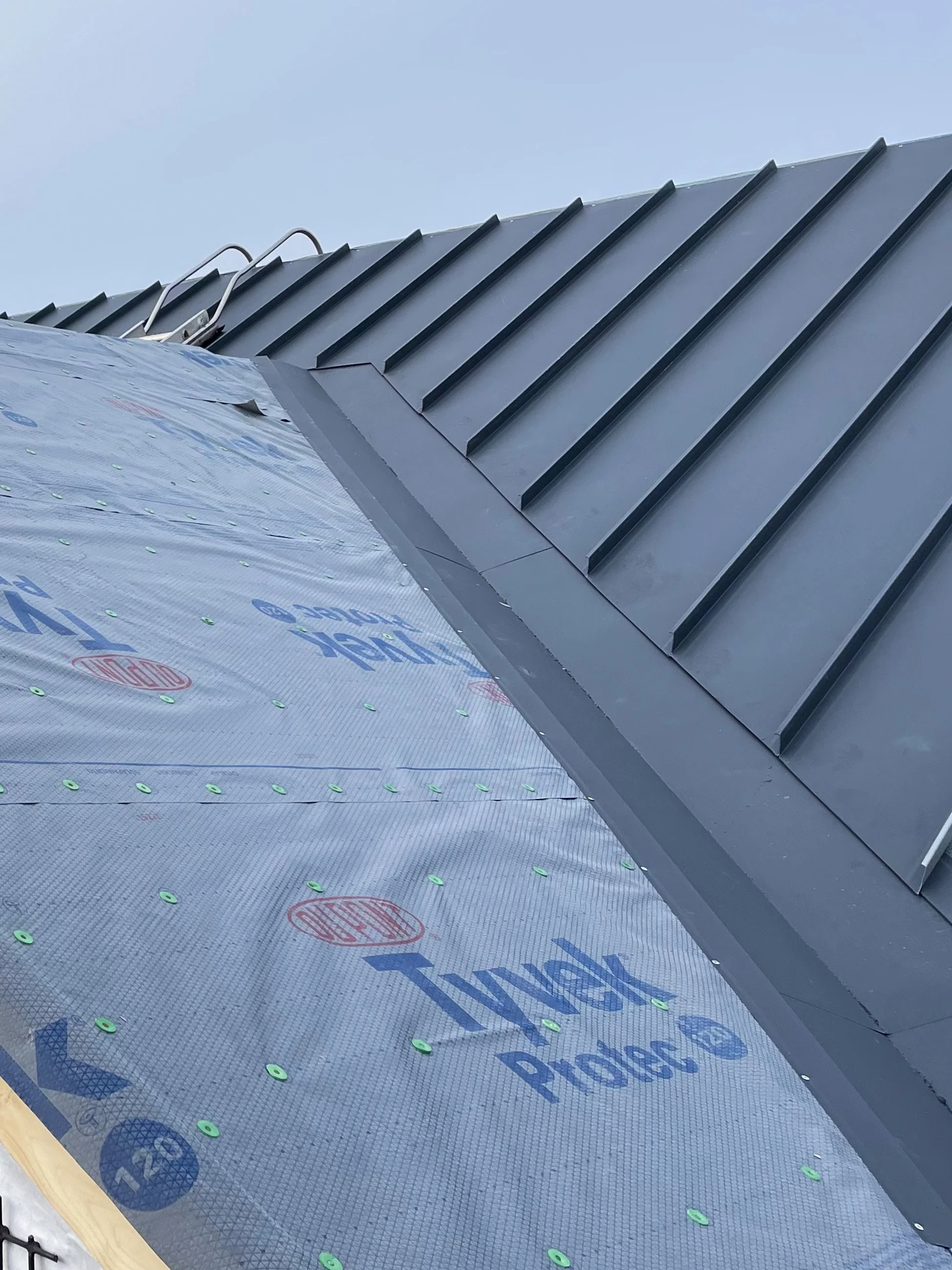 Close-up view of a metal roof under construction, showing roofing underlayment with branding and the metal roofing panels being installed along the roof ridge.