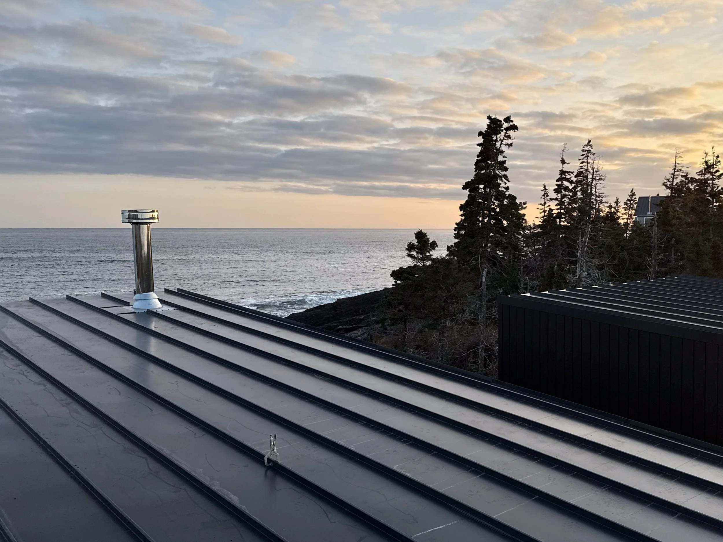 View of a metal roof with a chimney, overlooking an ocean with a rocky shoreline and a cluster of tall pine trees. The sky is partly cloudy with the sun setting or rising, creating a warm glow.