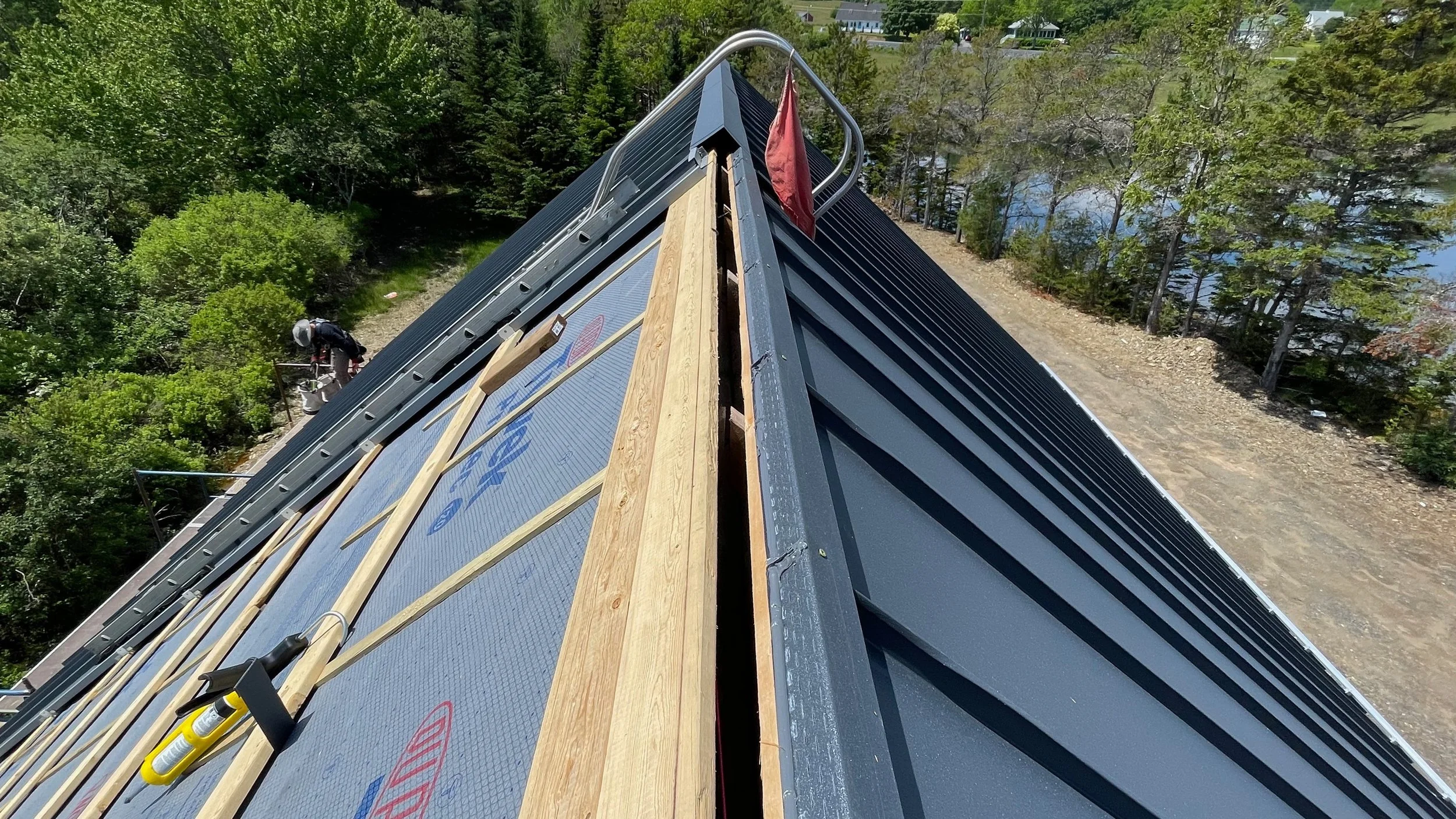 A house roof under construction with black metal roofing panels, wooden battens, and construction tools, surrounded by trees and a dirt road, viewed from the peak of the roof.