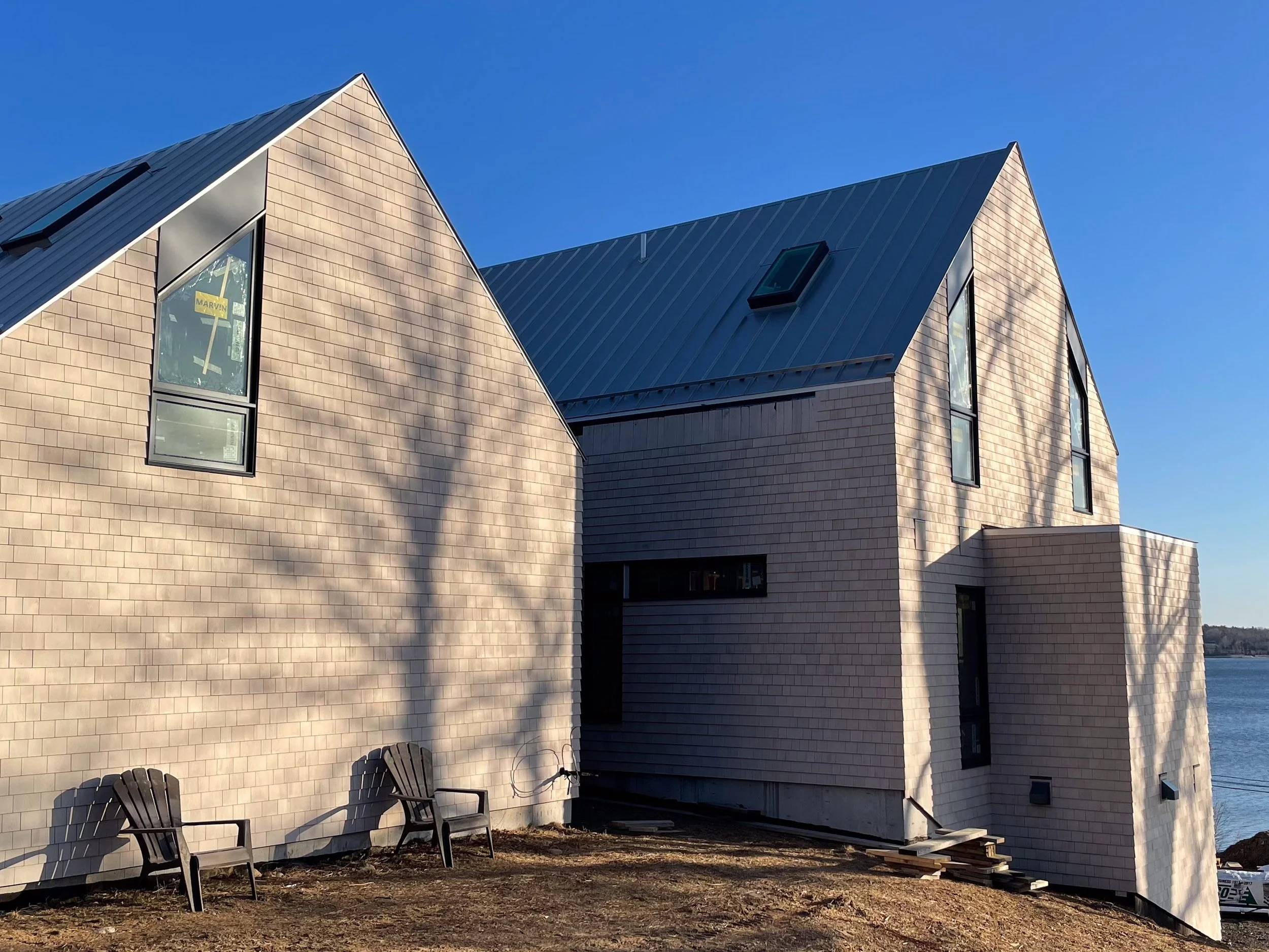 Modern house with beige brick exterior, sloped metal roof, and large windows near a body of water. Two black chairs are outside on dirt ground.
