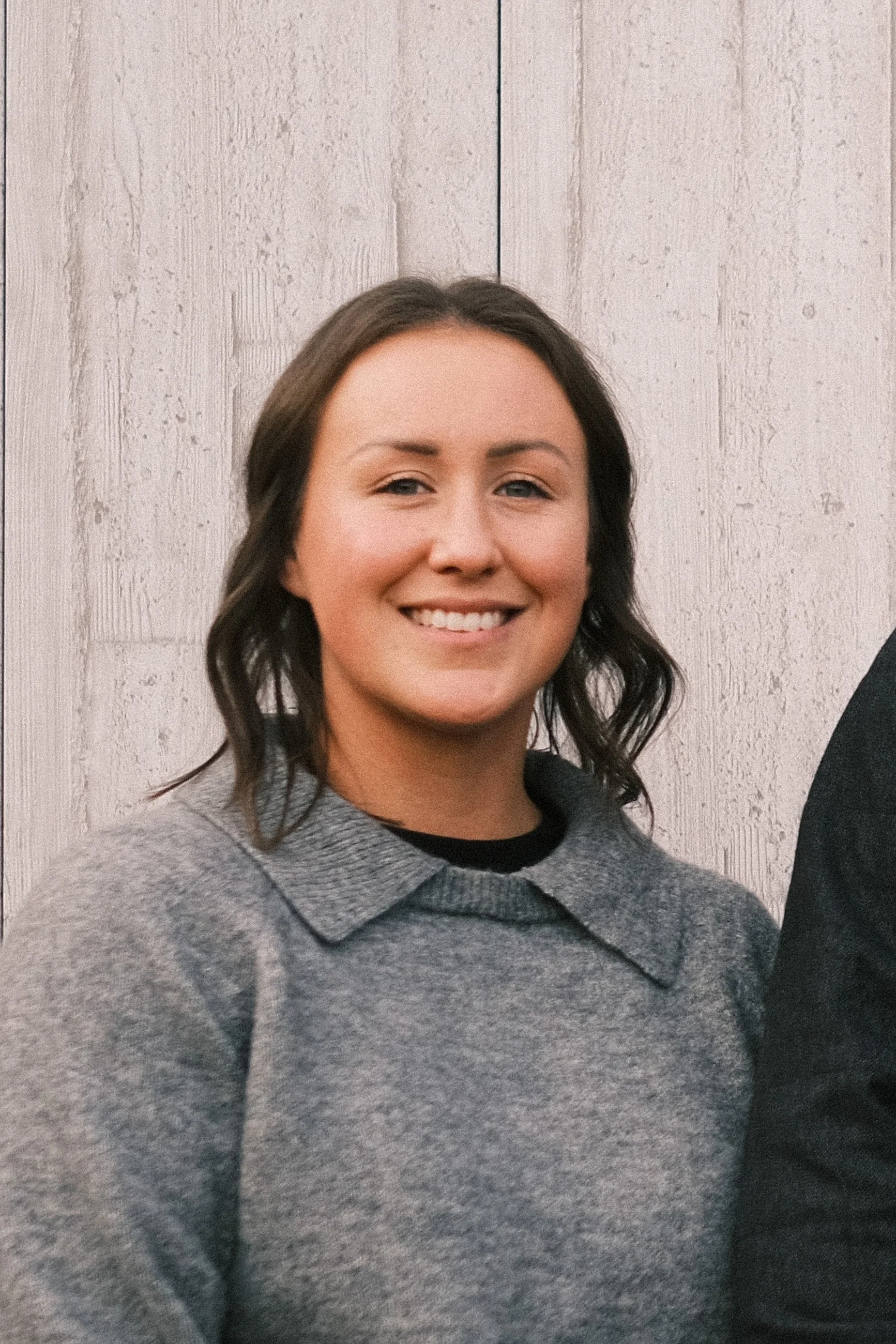 A smiling woman with shoulder-length brown hair wearing a gray sweater in front of a light wooden background.