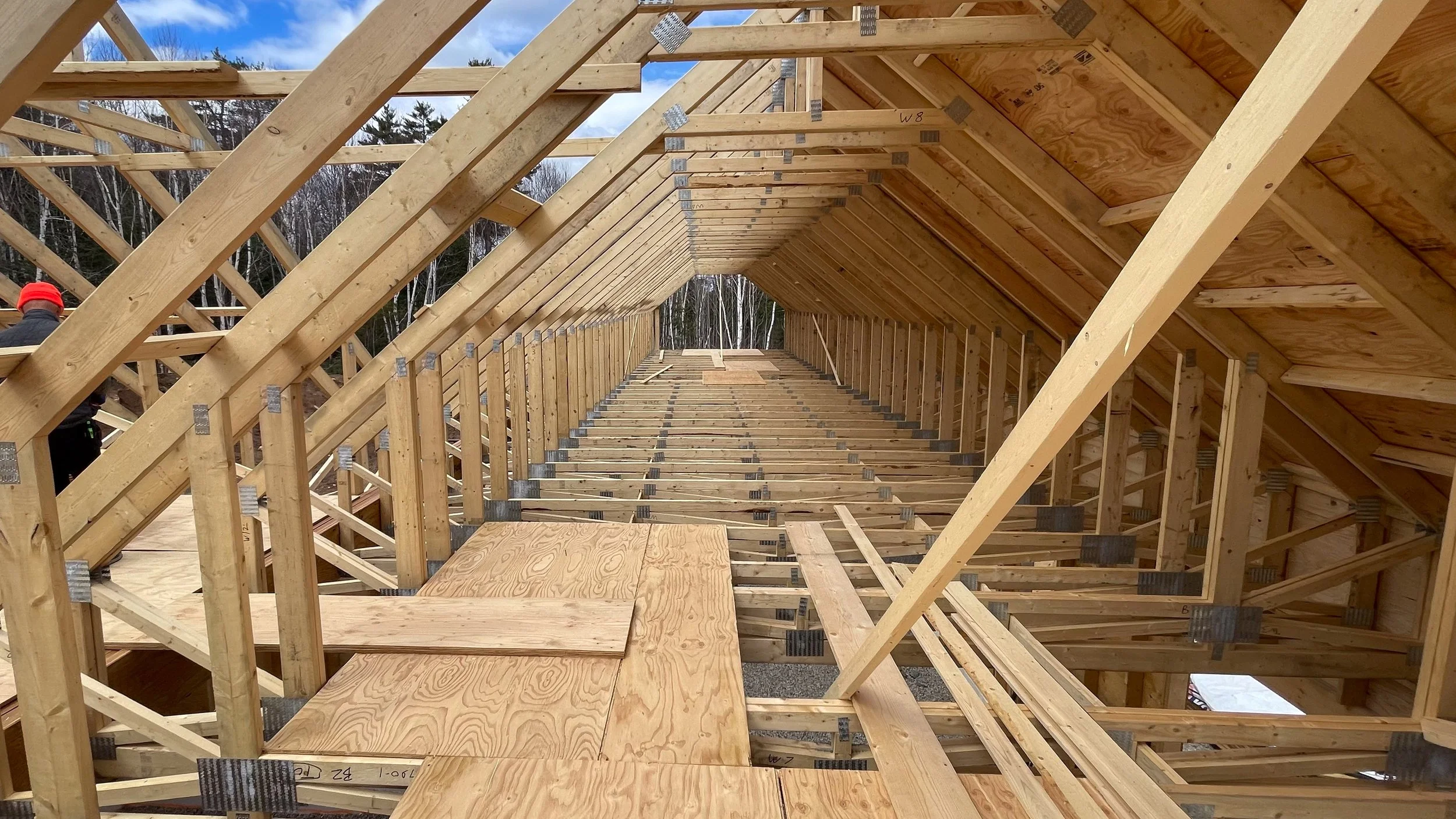 View of a building under construction showing wooden framing and roof trusses, with a worker wearing a red hard hat visible on the left side.