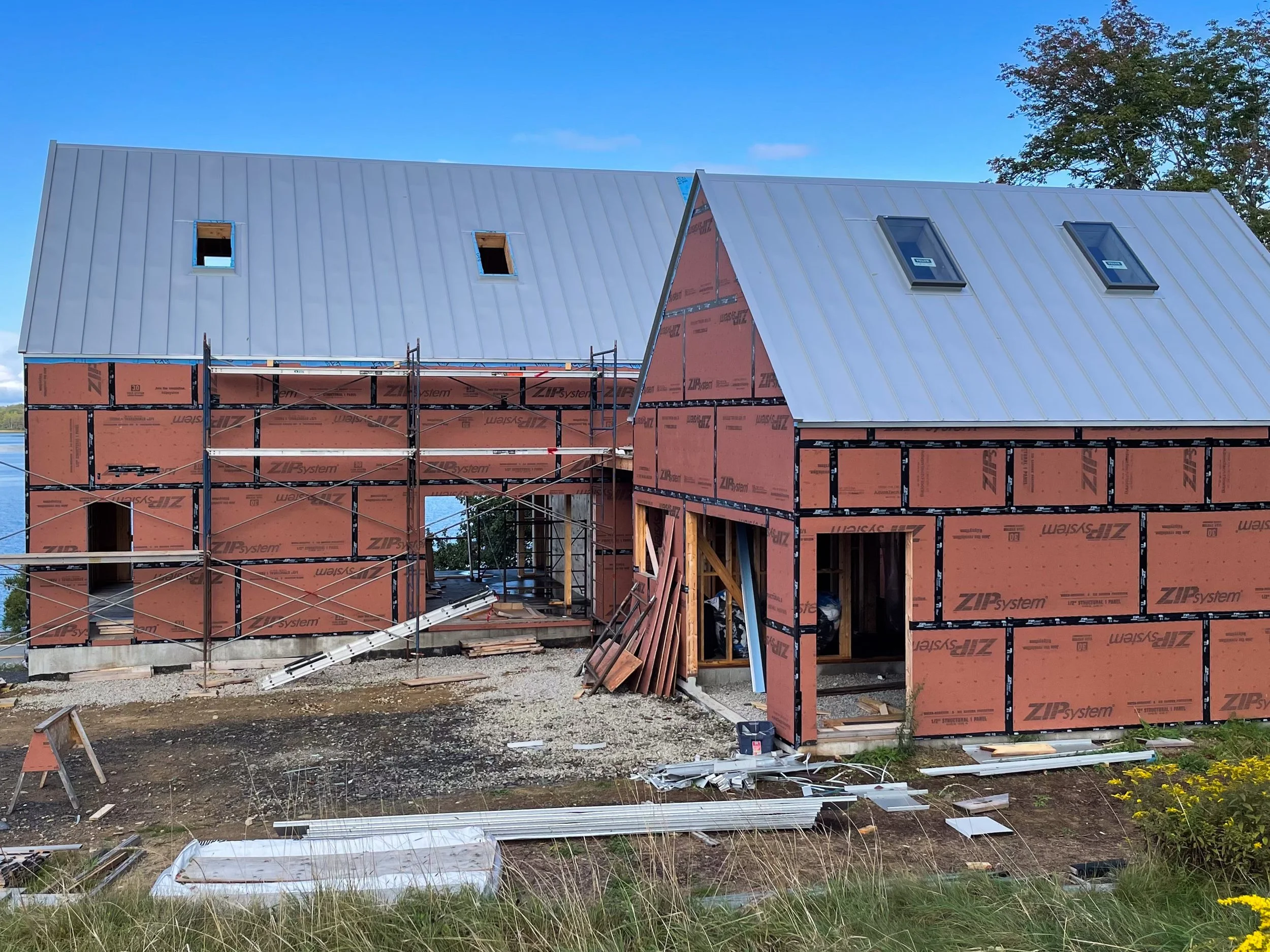 Two houses under construction with red exterior sheathing, scaffolding around the left house, and a metal roof on both houses. Small windows installed on the roof of each house. Construction materials scattered on the ground in front, with trees and 