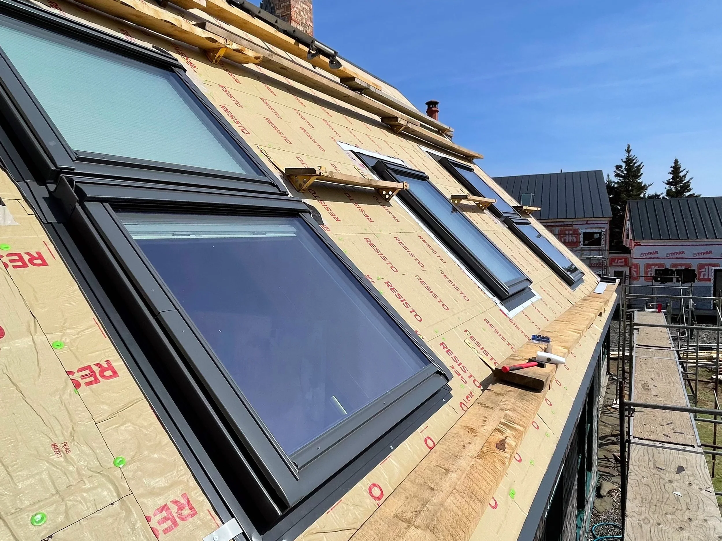 Roof under construction with multiple installed windows, scaffolding on the side, and bright blue sky in the background.