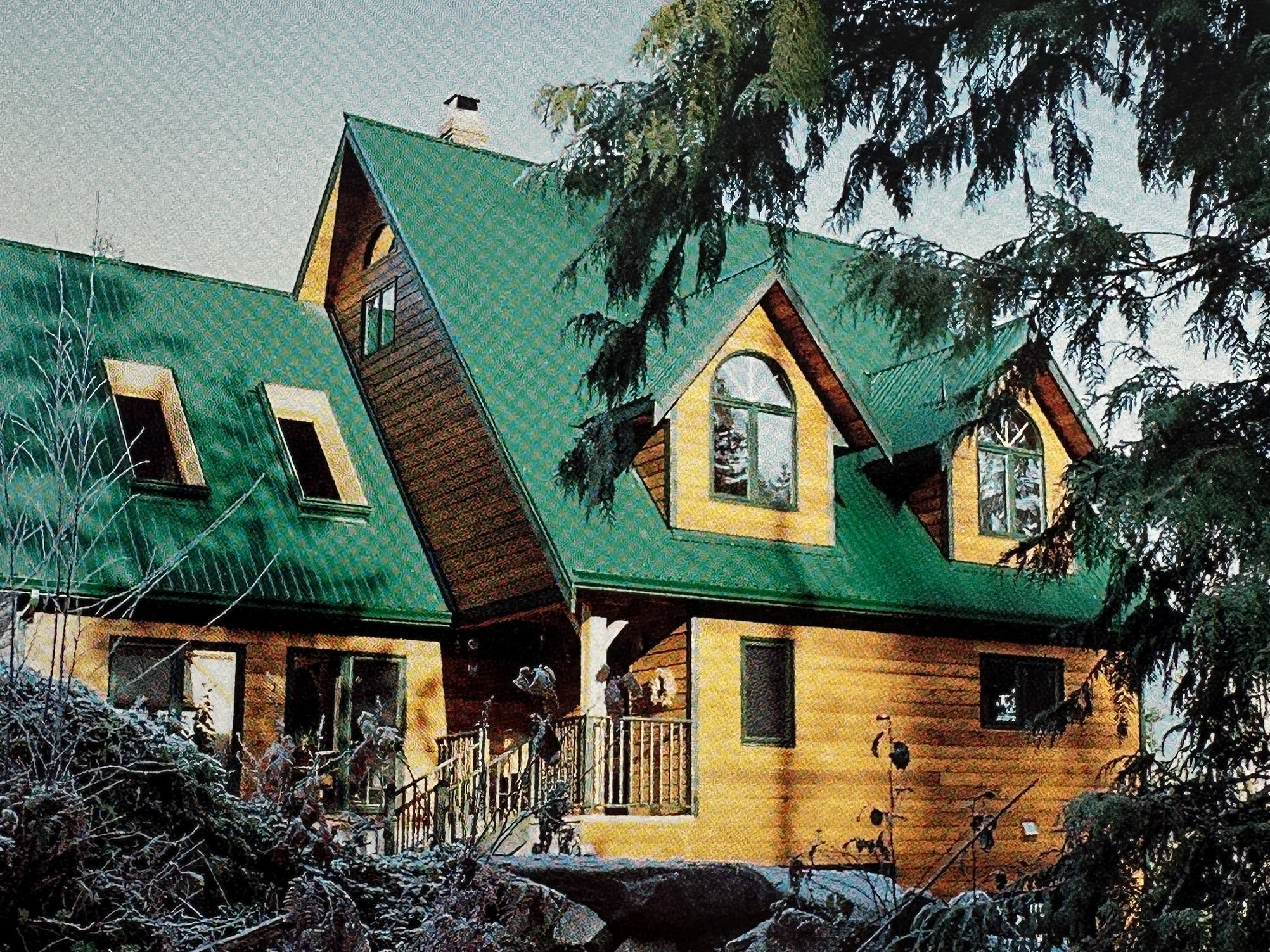 A large house with a green metal roof and yellow wooden exterior, surrounded by trees and rocks.