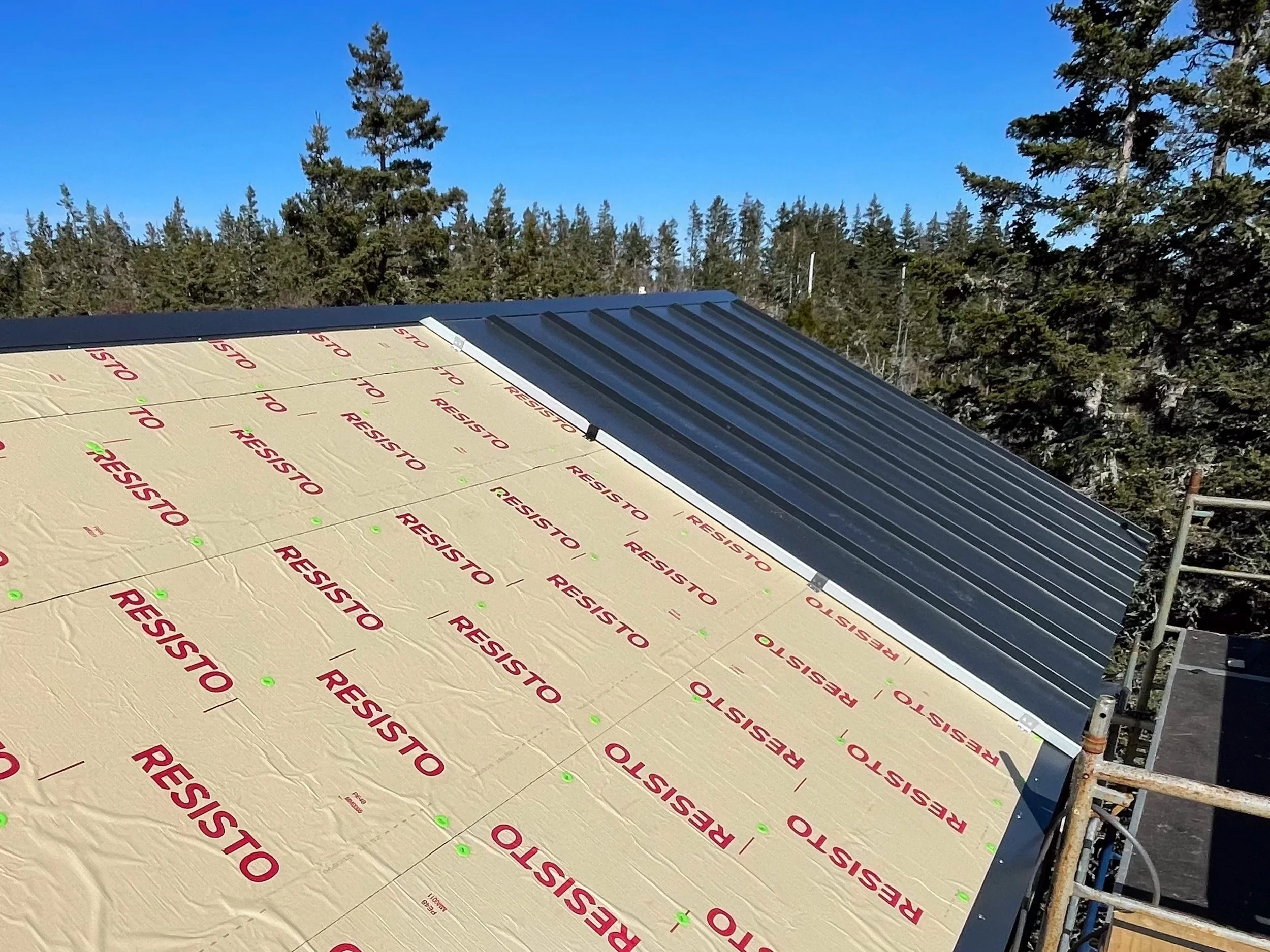 Construction roof with insulation material labeled 'Resisto' and metal roofing panels partially installed, surrounded by scaffolding, trees, and clear blue sky.