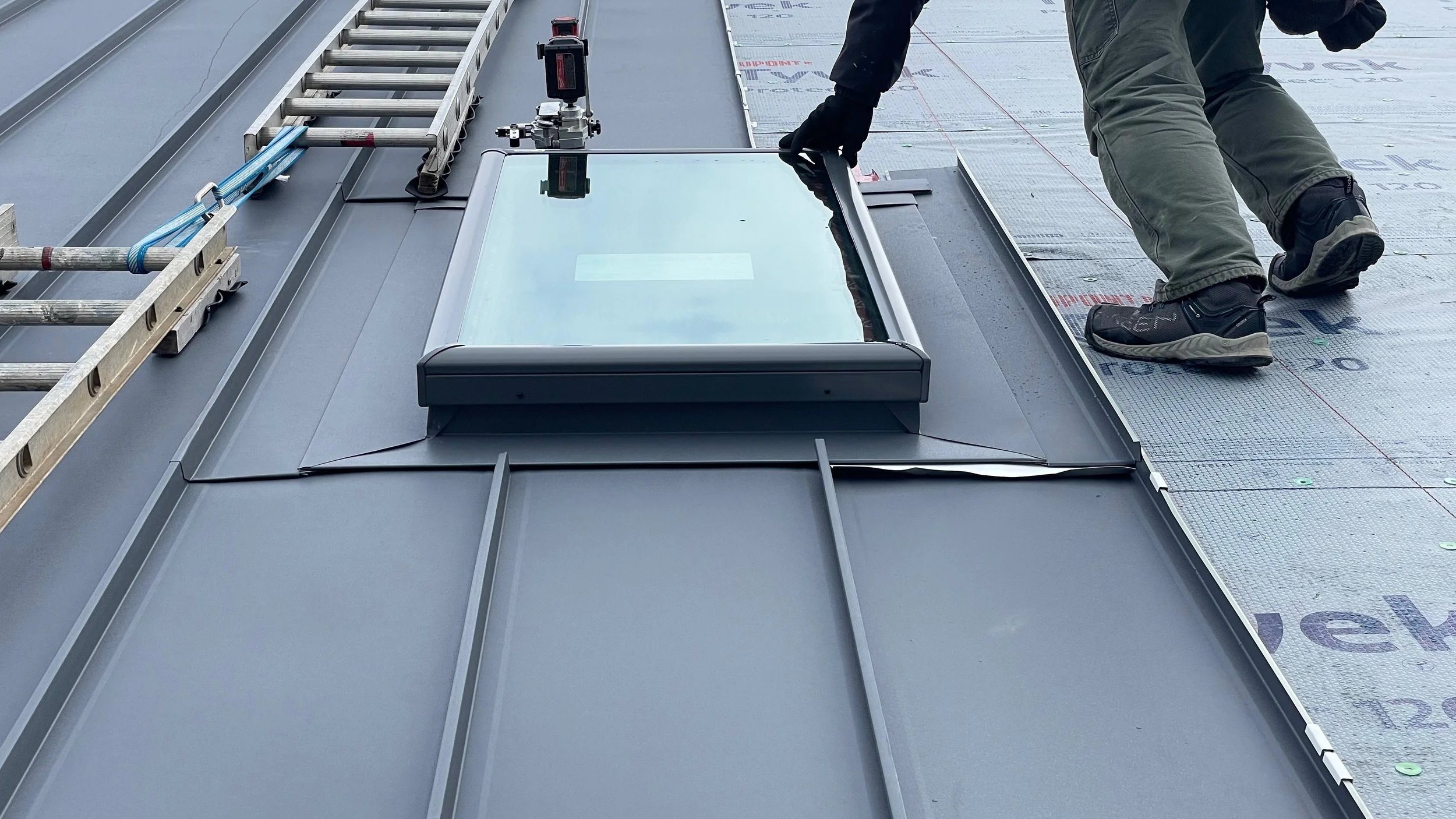 A worker installing a solar skylight on a metal roof, with tools and ladder nearby.