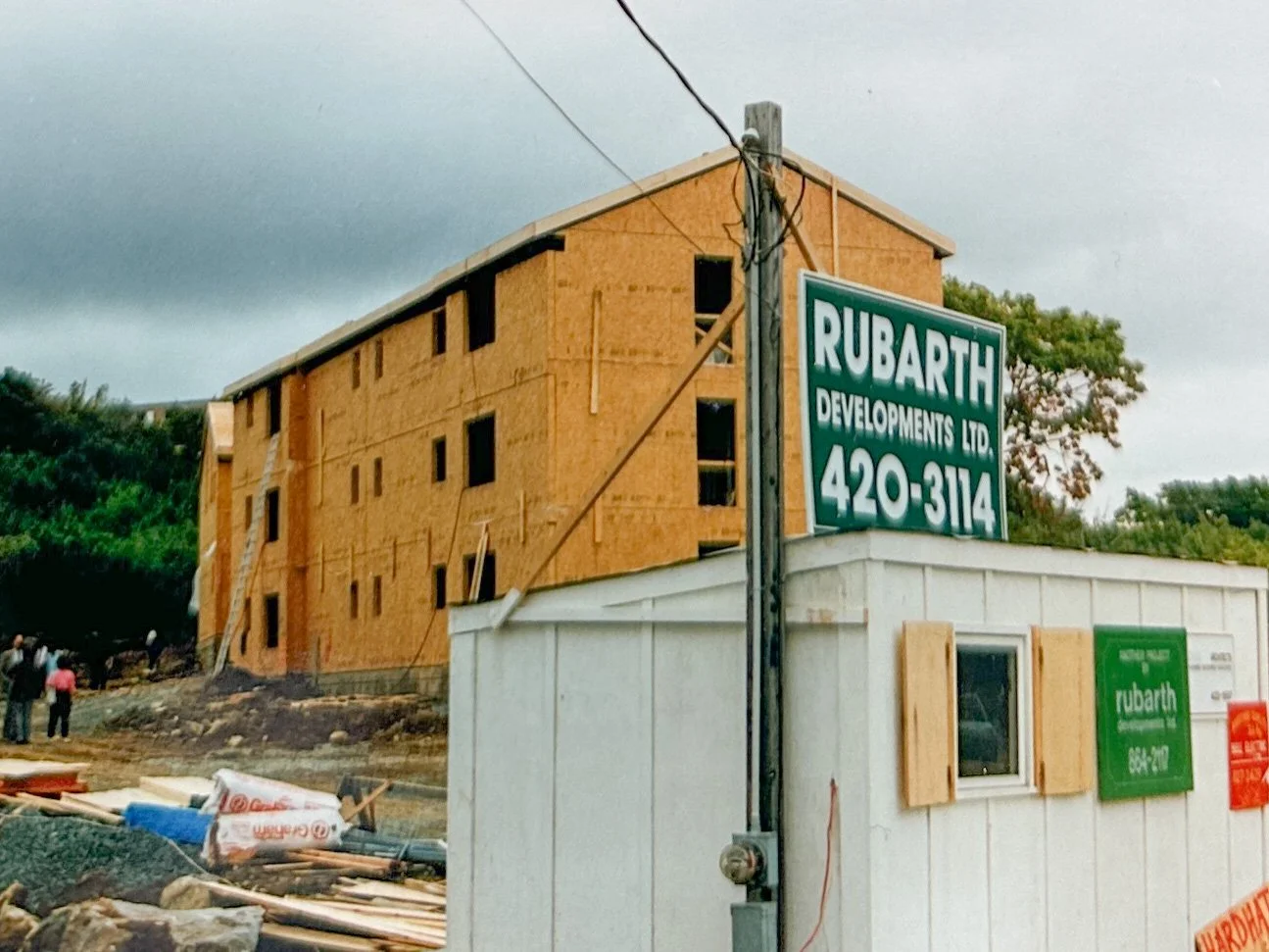 Construction site with a partially built wooden apartment building and a small white office or construction booth. Sign on pole displays 'Rubarth Developments Ltd 420-3114.' Overcast sky with trees in the background.