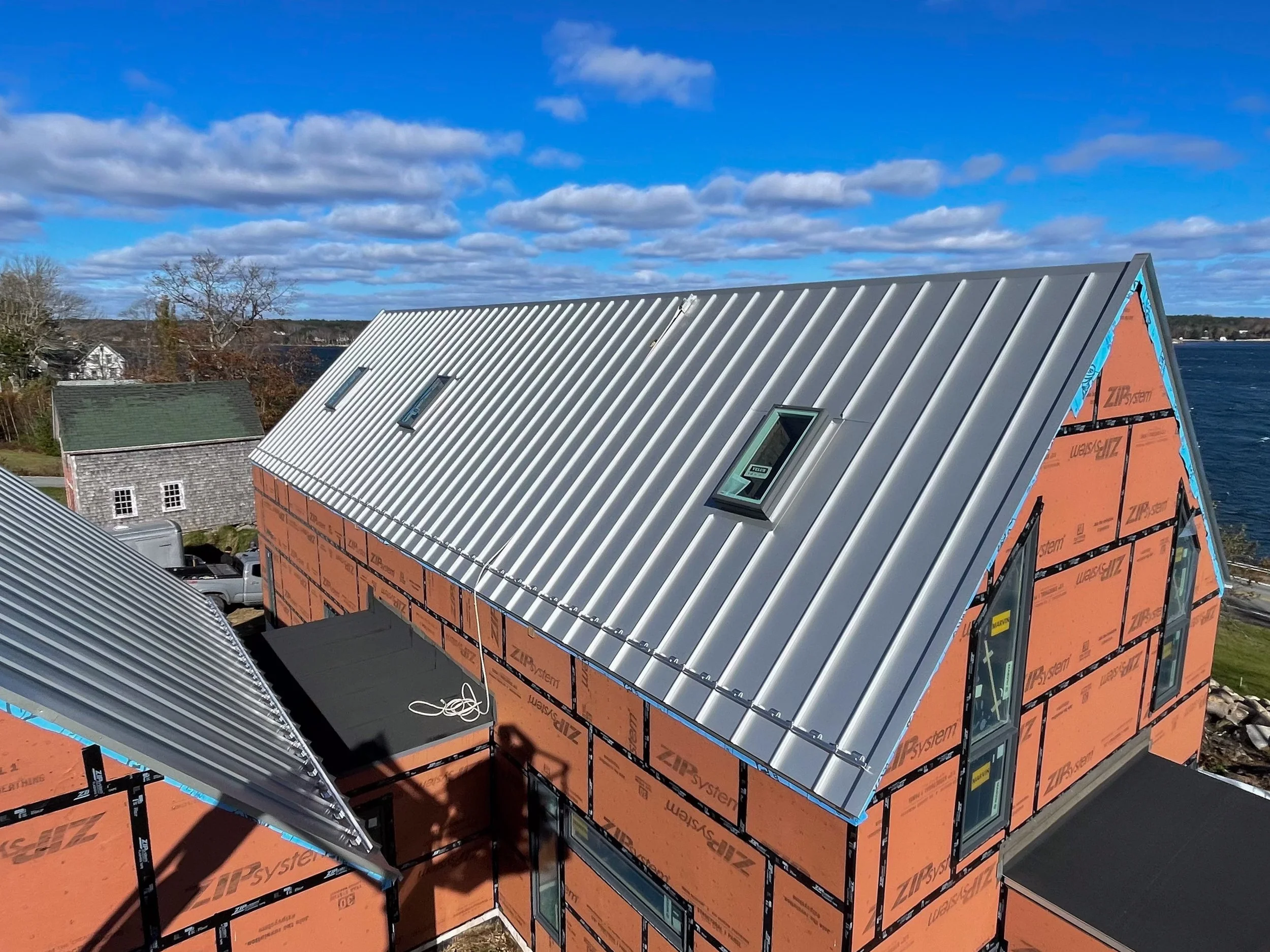 Under construction house with metal roof and windows, surrounding homes, water body, and blue sky with clouds.