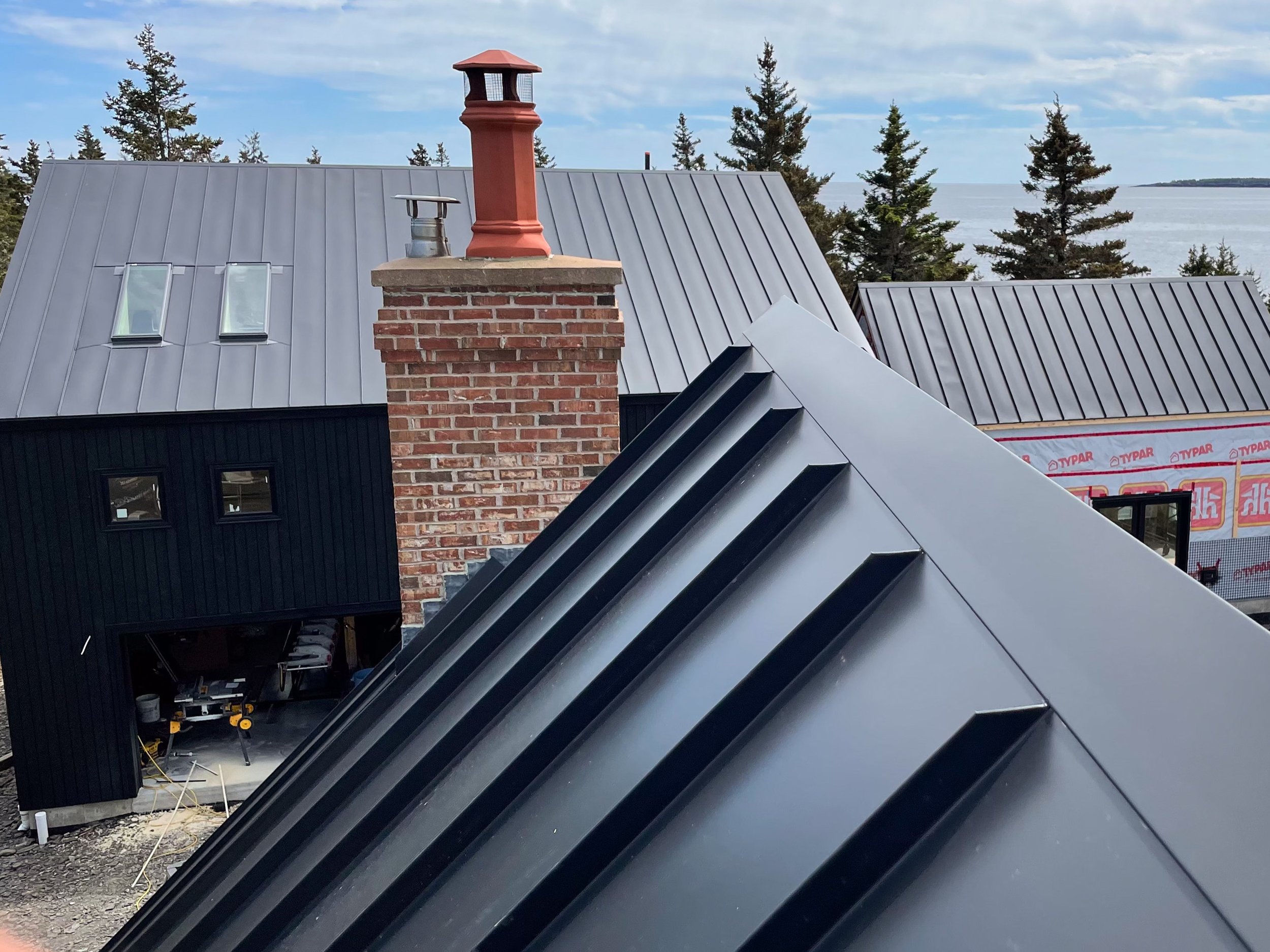 View of rooftops with metal roofing, brick chimney, and windows, overlooking a body of water and trees in the background.