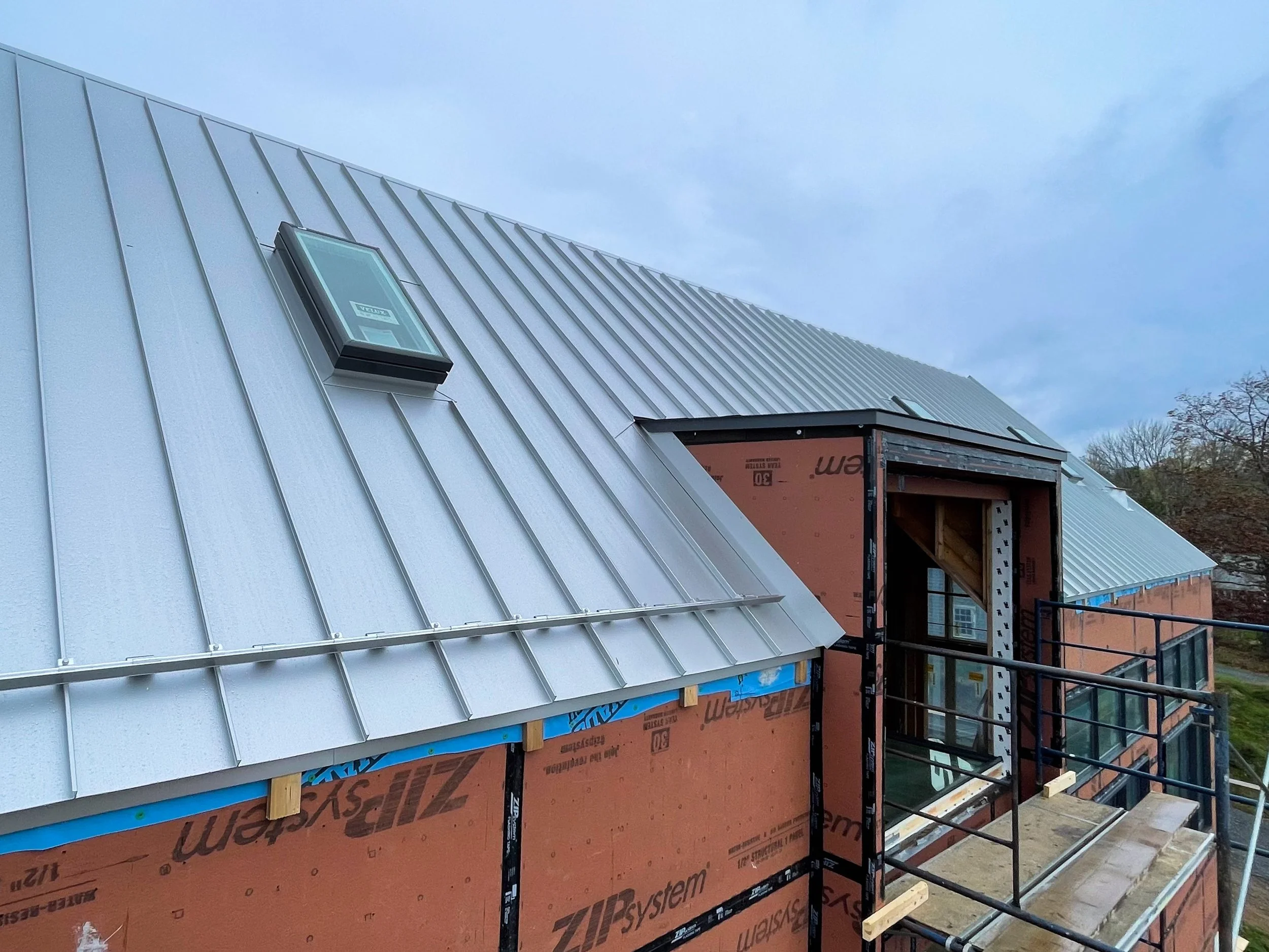 Construction site showing a house with a metal roof, a dormer under construction, and scaffolding, with cloudy sky in the background.