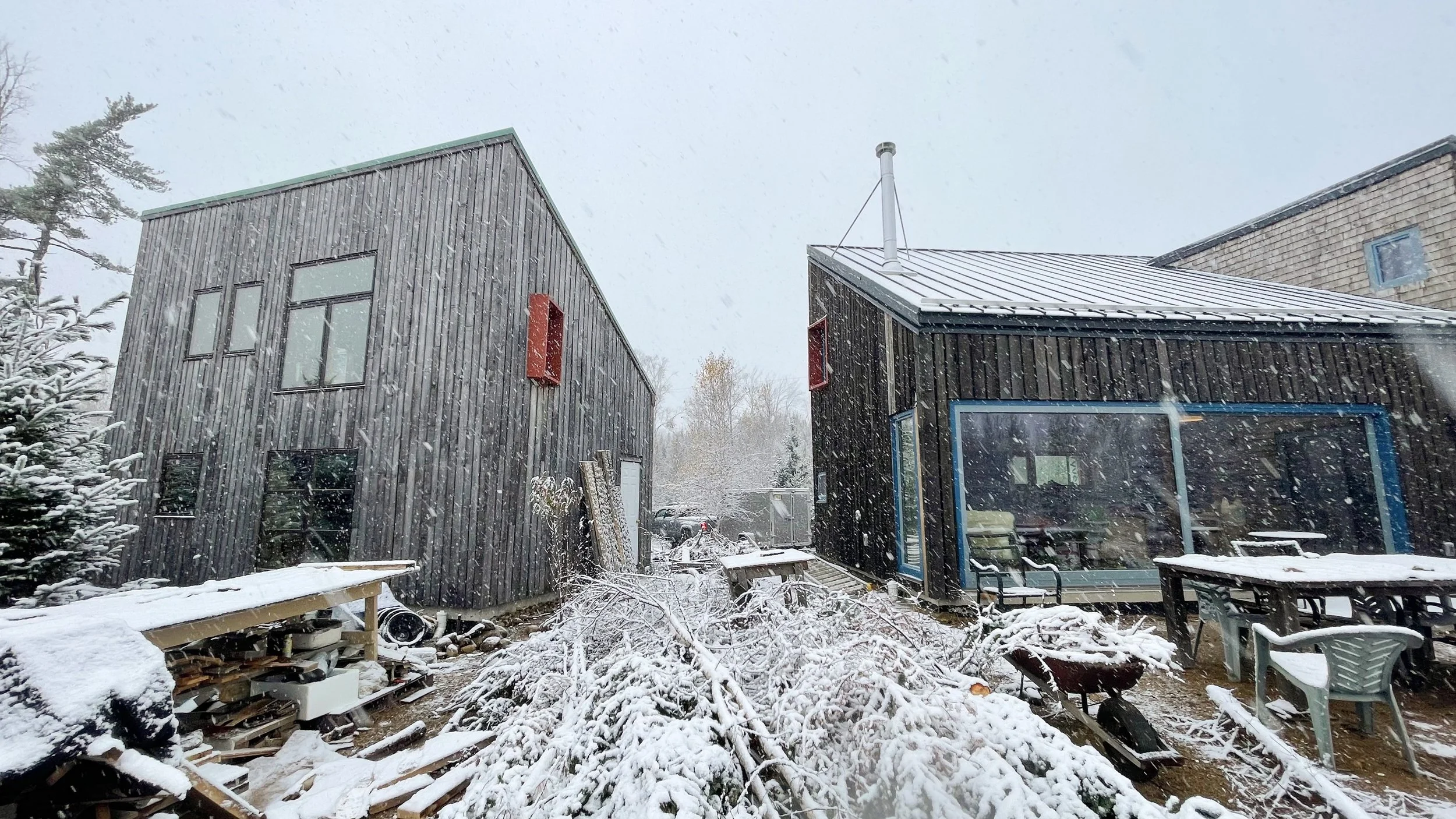 Snow falling in a backyard with two modern black wooden houses, outdoor furniture covered in snow, and a pile of snow-covered branches and debris in the foreground.