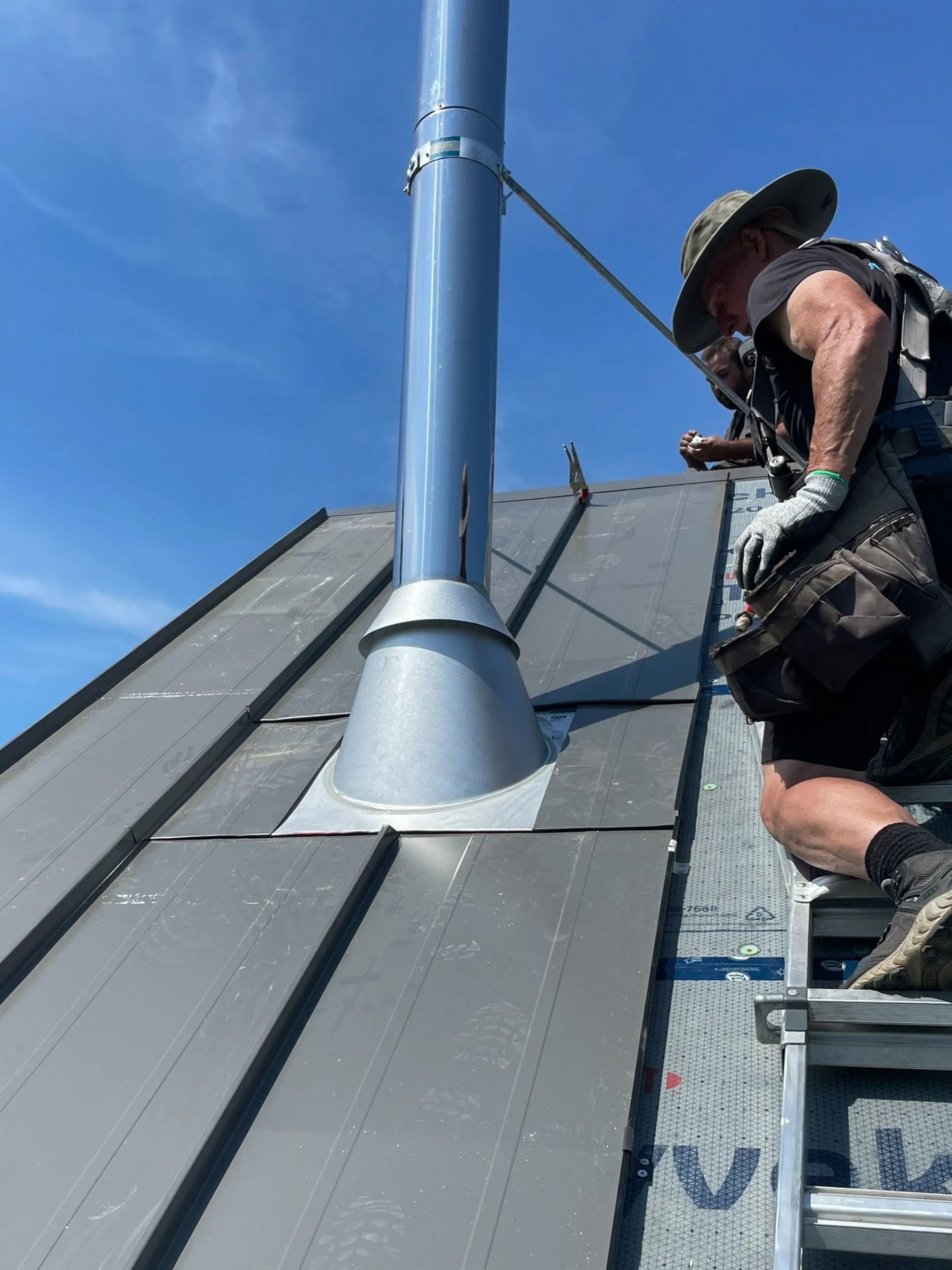 Worker installing or inspecting a metal vent pipe on a roof under a clear blue sky.