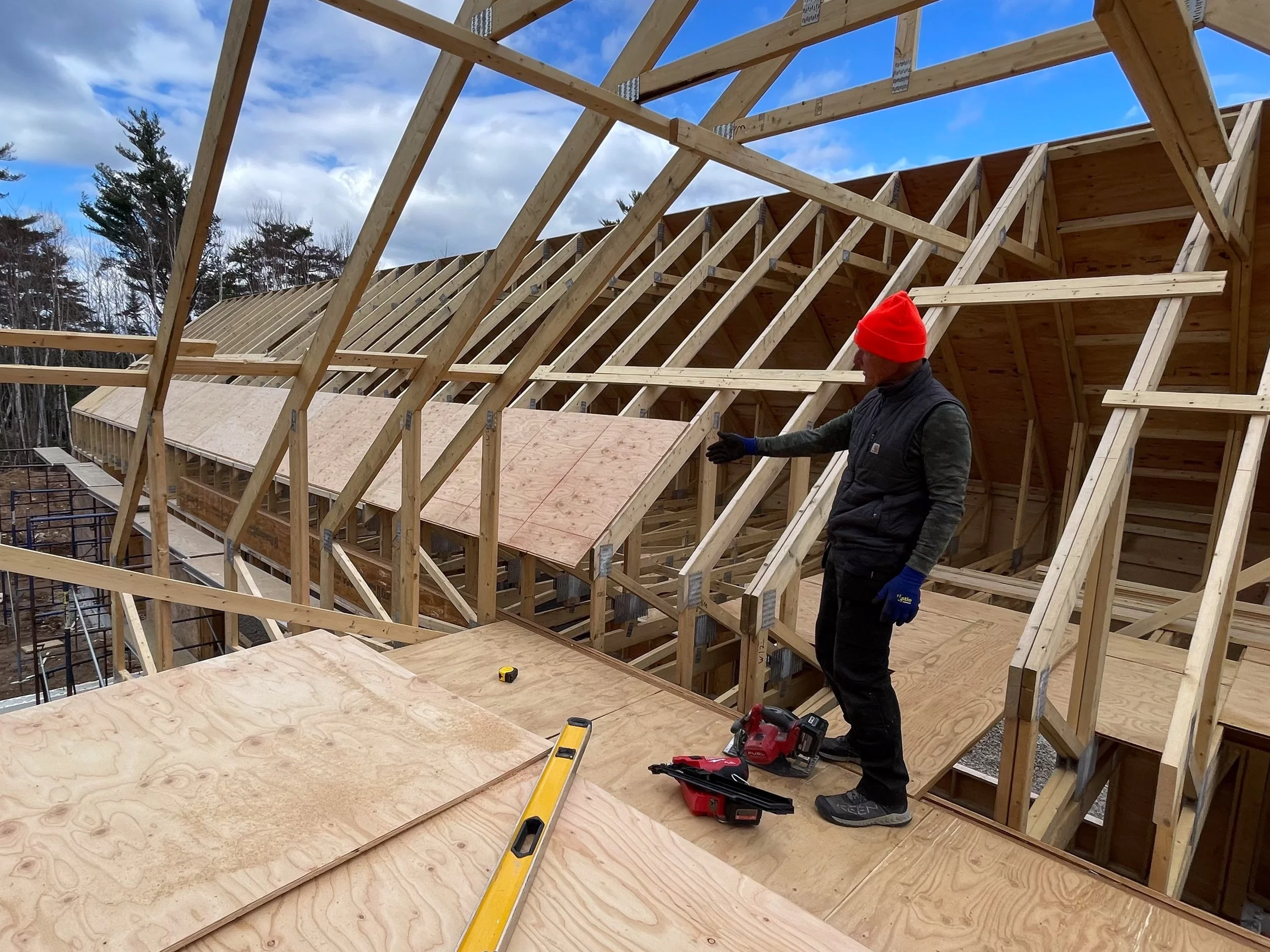 A construction worker wearing a red beanie, gloves, and a black vest is standing on a wooden platform at a building site. He is pointing at or inspecting the wooden roof framework of a house under construction, with a bright blue sky and some clouds above.