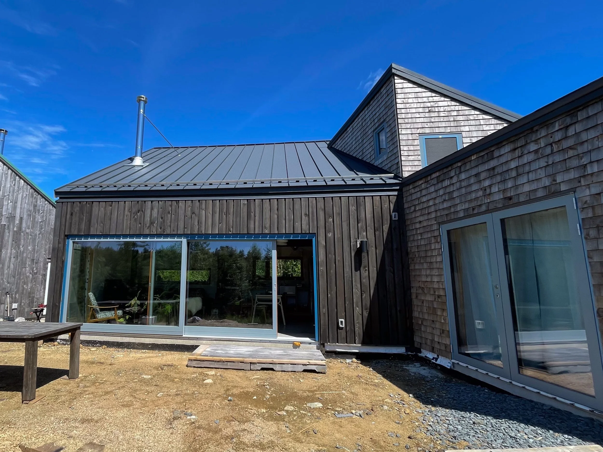 Exterior view of a modern house with dark wooden siding, large glass sliding doors, and a metal roof. Clear blue sky in the background.