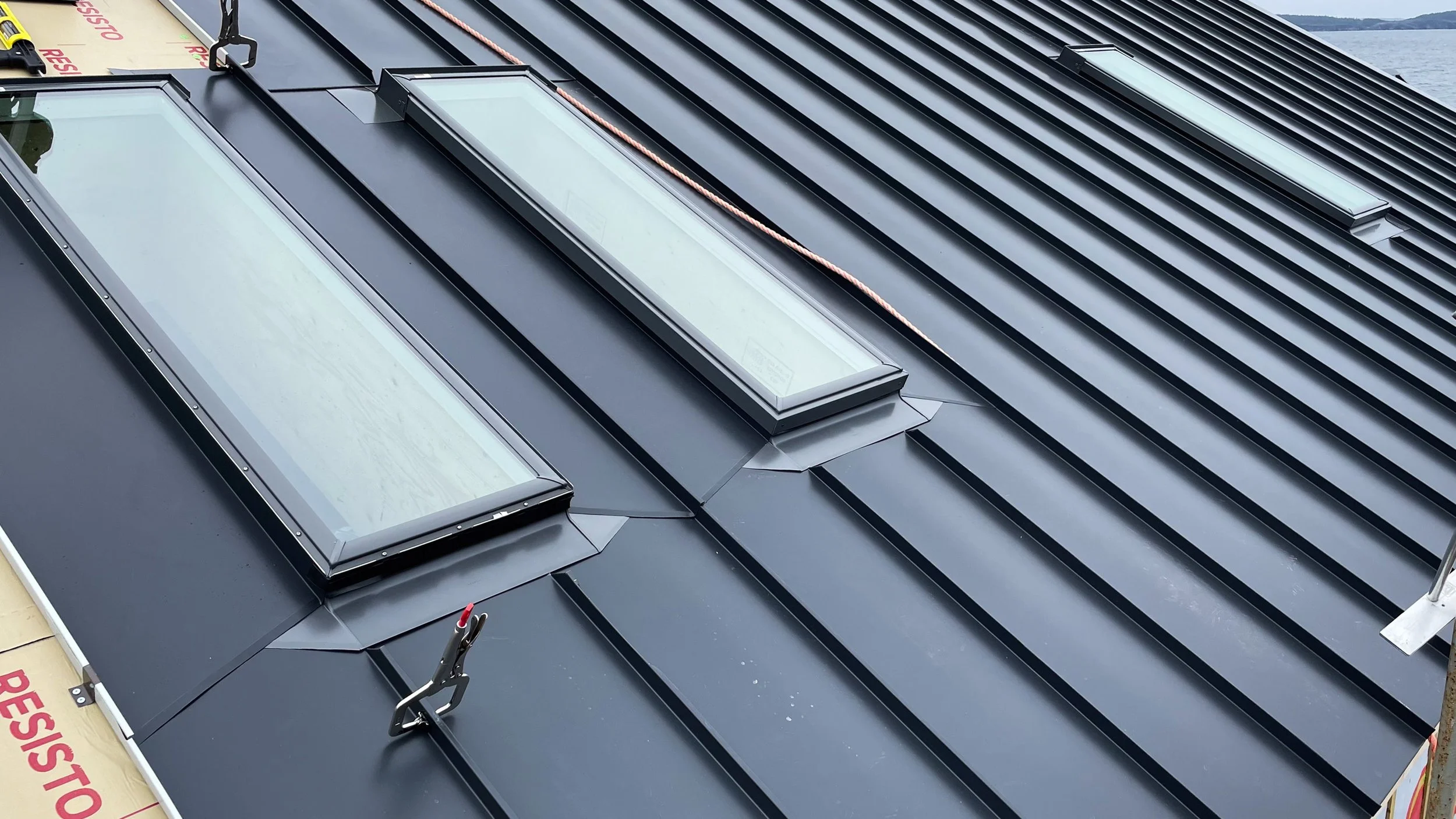 Metal roof with three skylights and a clothes clip attached to a cable, overlooking a body of water.
