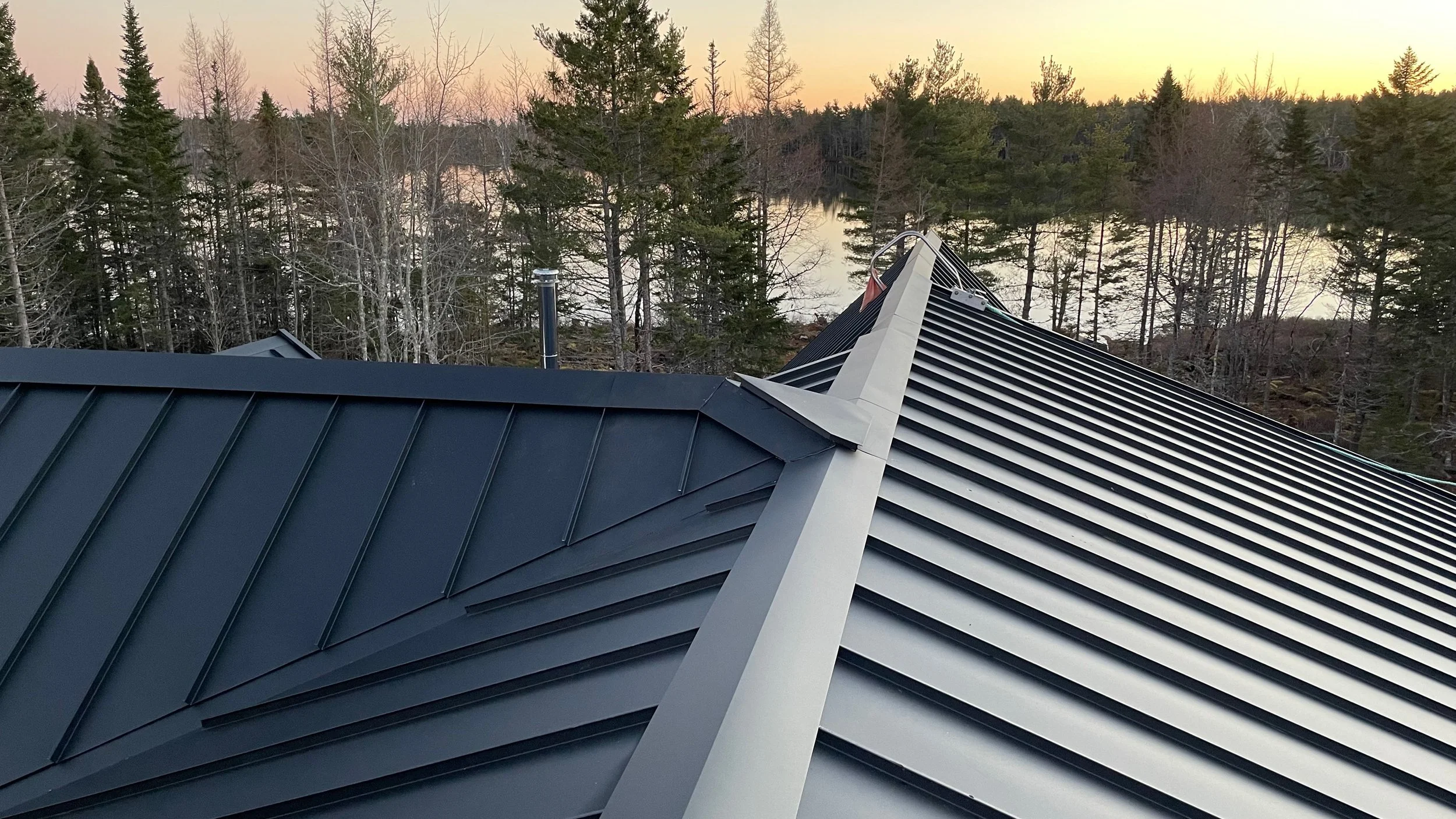 A metal roof with a gable design, surrounded by trees, overlooking a body of water and a sunset sky.