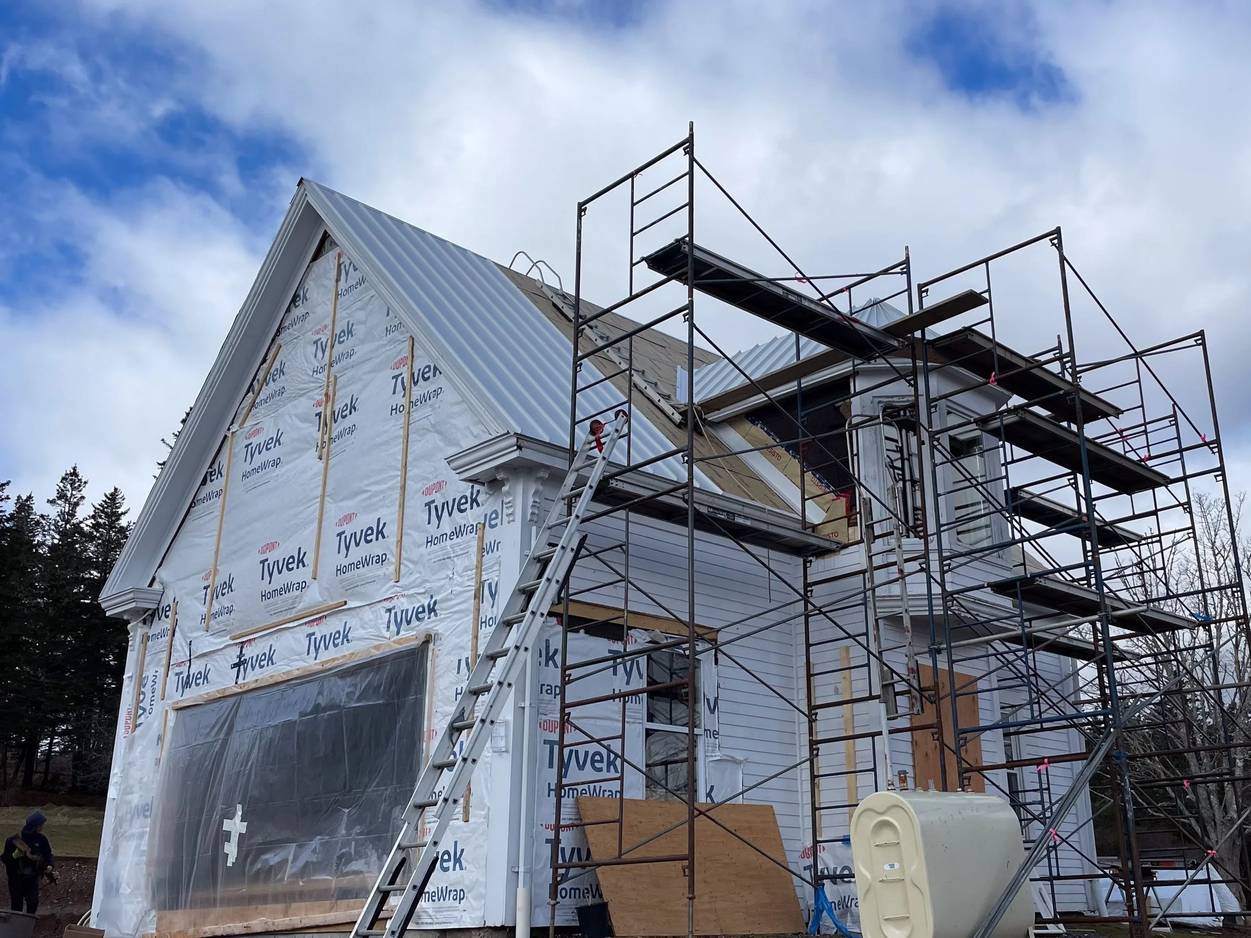 Partial view of a house under construction, covered in Tyvek house wrap, with scaffolding around it, and a person working on the left side.