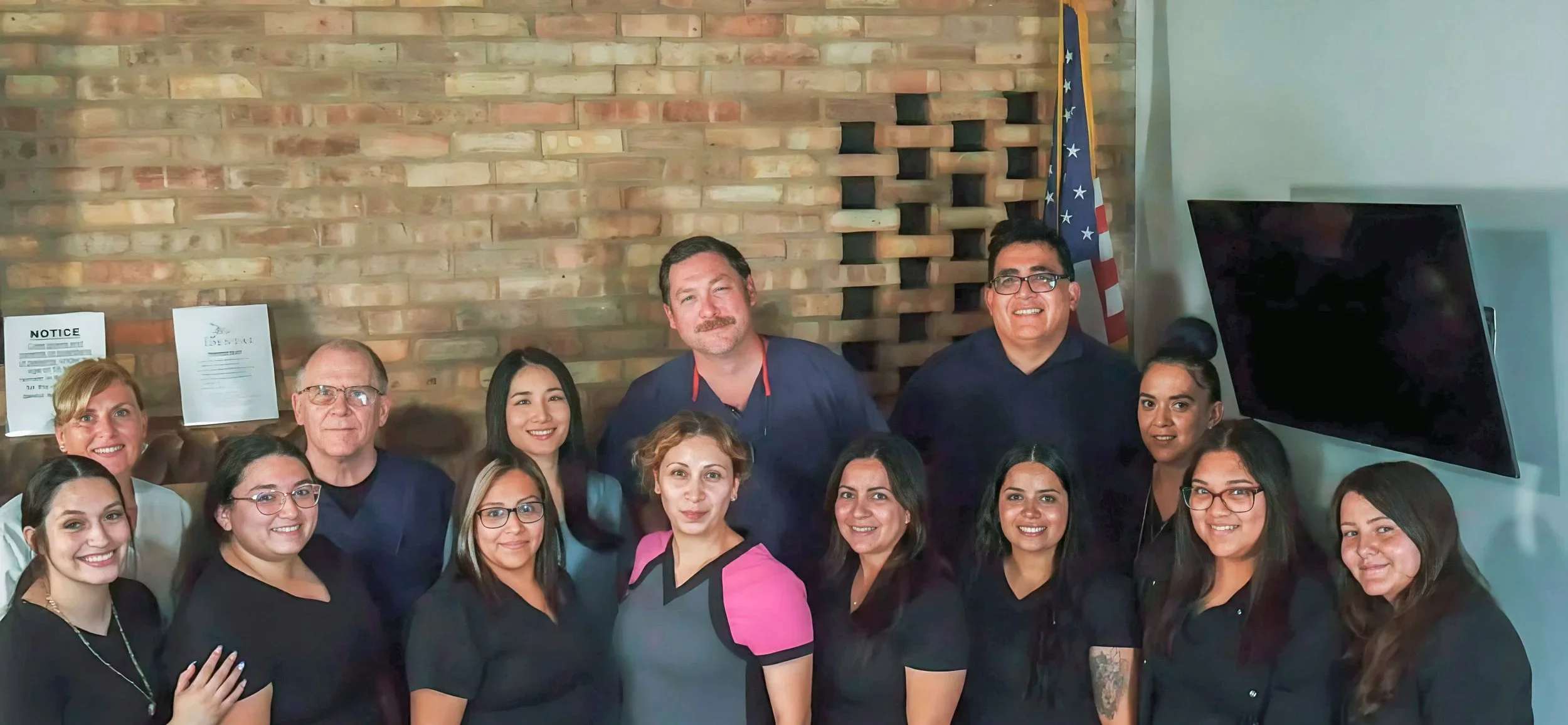 Group of thirteen diverse people posing indoors, smiling, with a brick wall and American flag in the background.