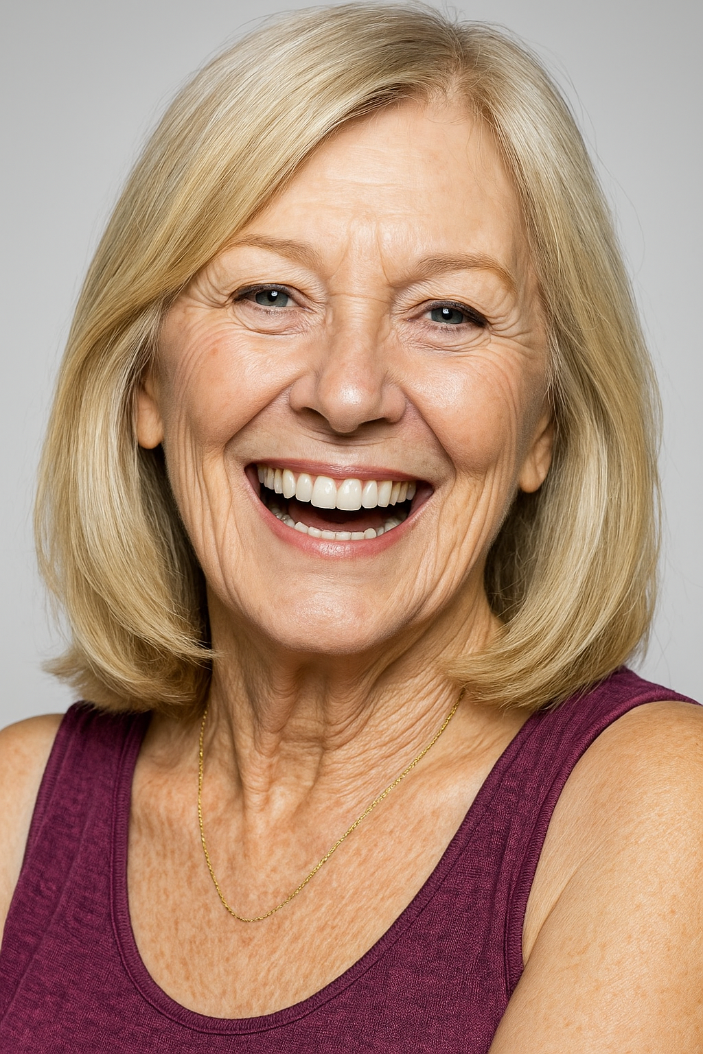 Close-up of a smiling elderly woman with shoulder-length blonde hair, wearing a sleeveless purple top and a gold necklace, against a plain light grey background.